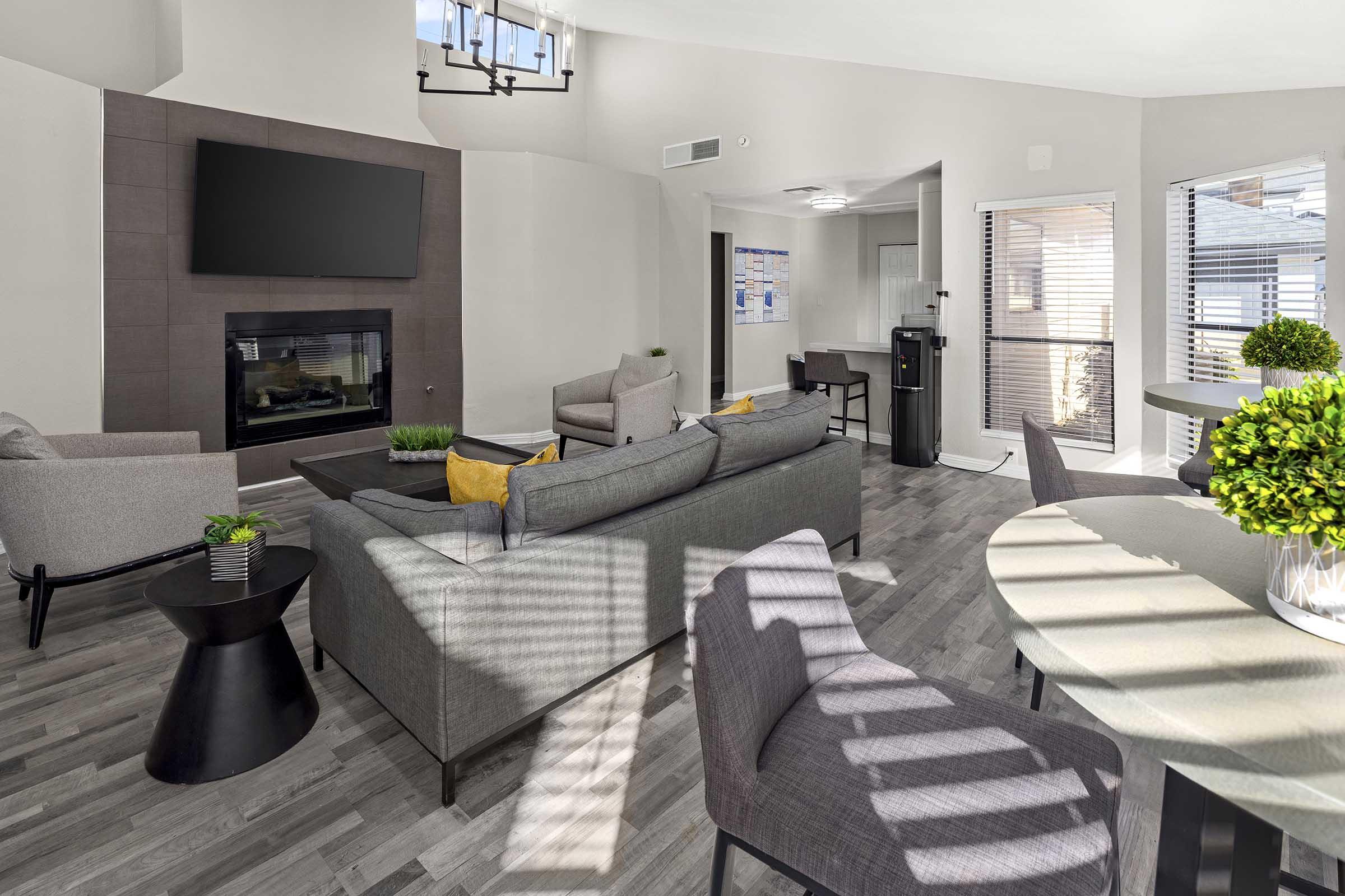 A modern living area featuring a gray sectional sofa, armchairs, and a round dining table. A flat-screen TV is mounted on the wall above a fireplace. Bright natural light streams through windows, creating a warm atmosphere. Minimalist decor with potted plants adds to the inviting space.