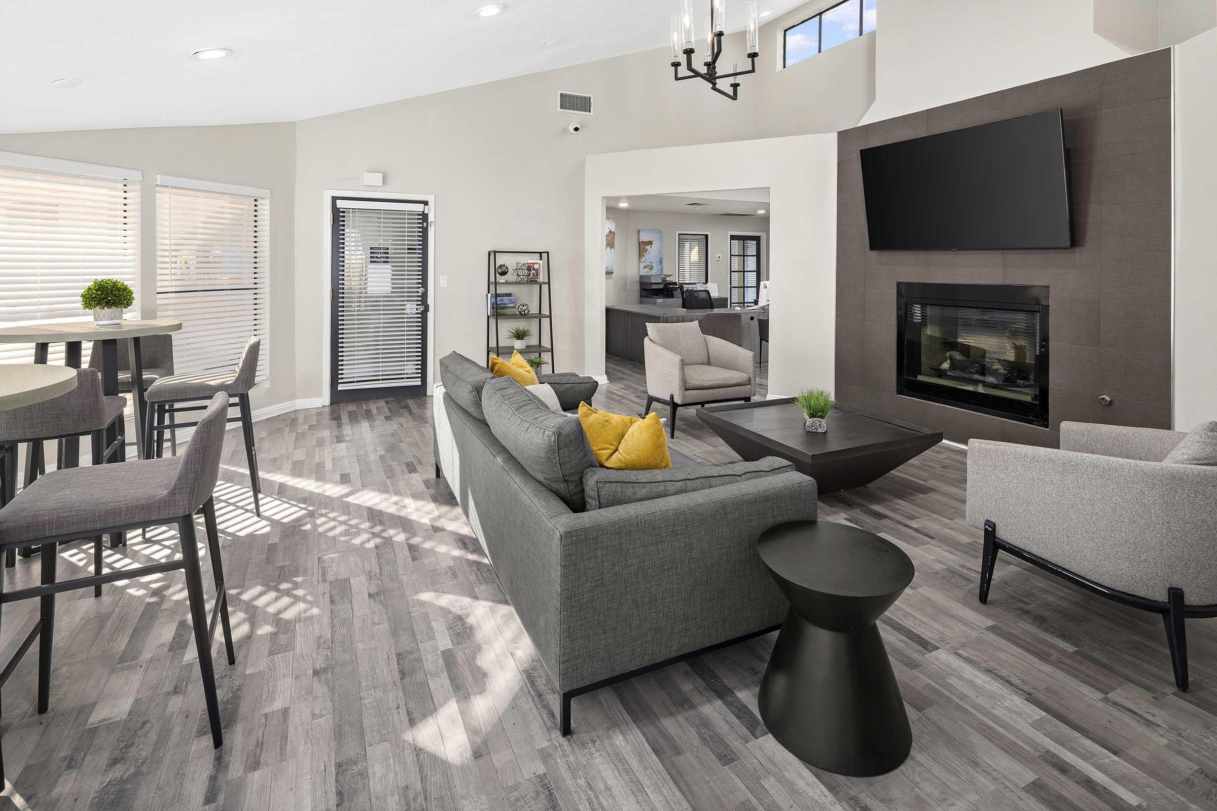 A modern living space featuring a cozy seating area with a gray sofa adorned with yellow pillows, a coffee table, and a stylish black side table. Large windows provide natural light, and there's a fireplace and a television mounted on the wall. Nearby, a dining area includes high-backed stools around a circular table.