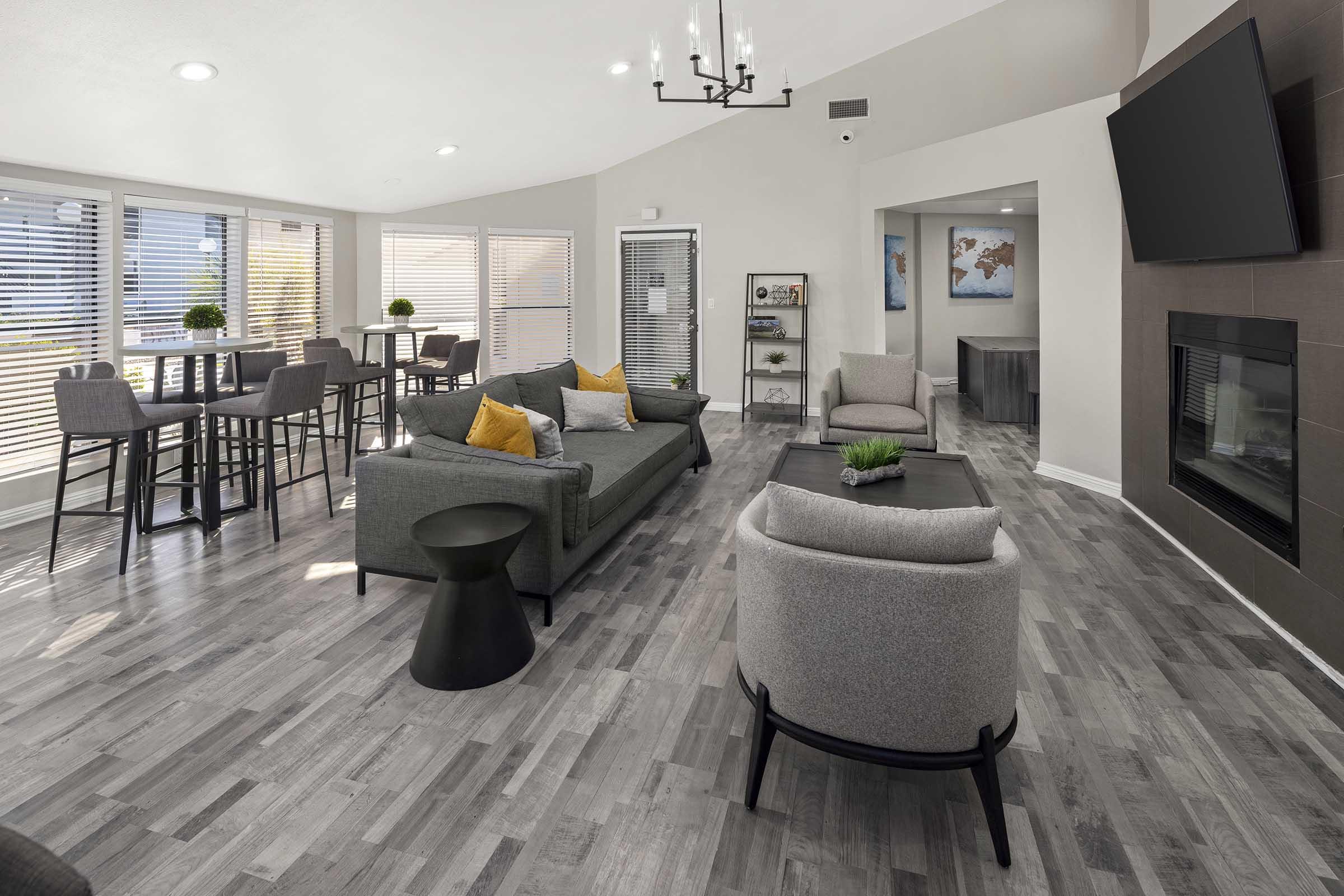 A modern living room featuring a cozy green sofa with orange pillows, light gray armchairs, a coffee table, and a small black side table. Natural light pours in through large windows with blinds. There's a fireplace and a wall-mounted TV, creating an inviting atmosphere. The decor is contemporary and stylish.