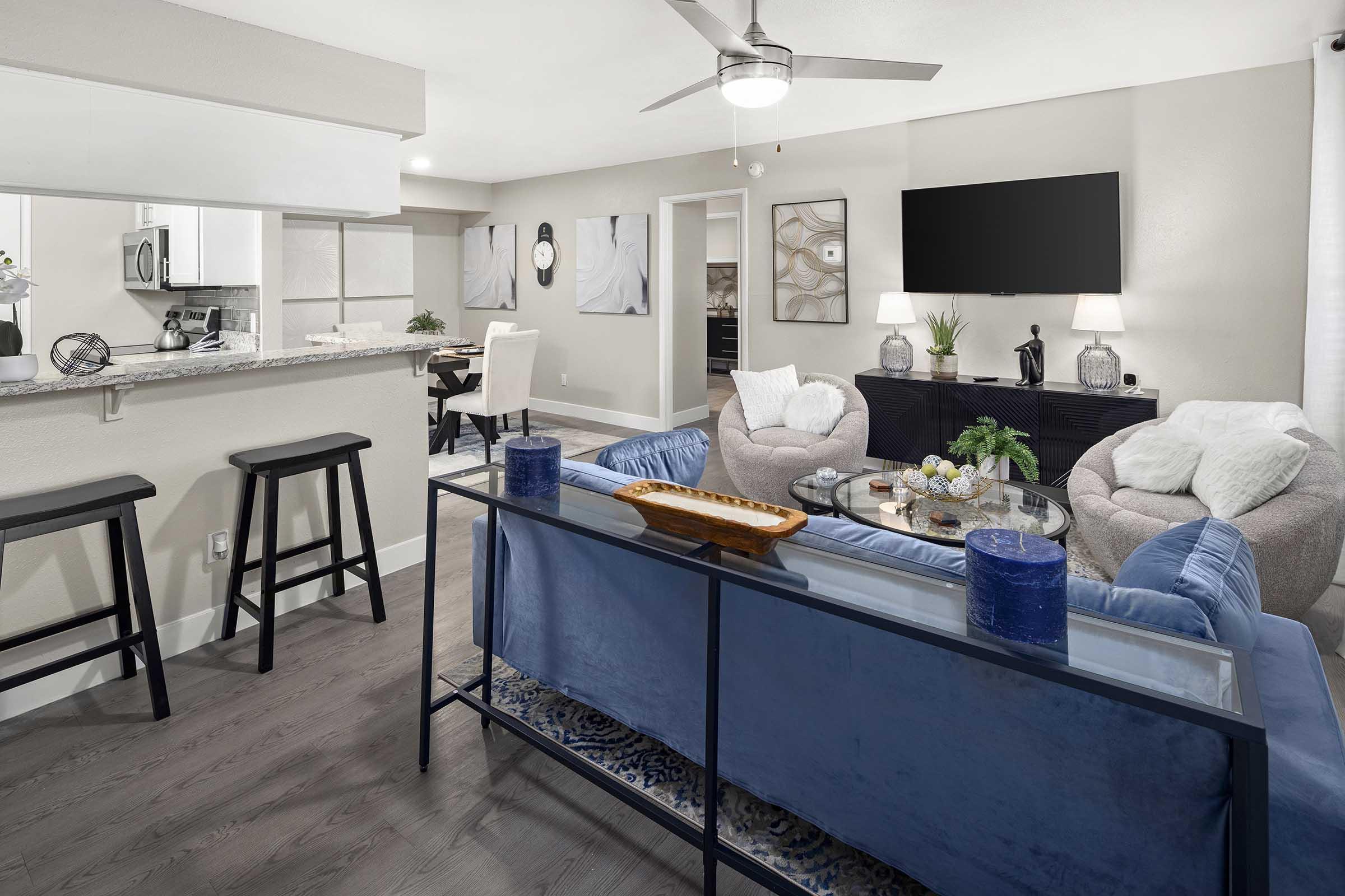 Modern living room with stylish furniture, including a blue couch and glass coffee table. Features a neutral color palette, contemporary art on the walls, and a flat-screen TV mounted above a black console. A dining area is visible, with a bar counter and stools in the background, creating an open and inviting space.
