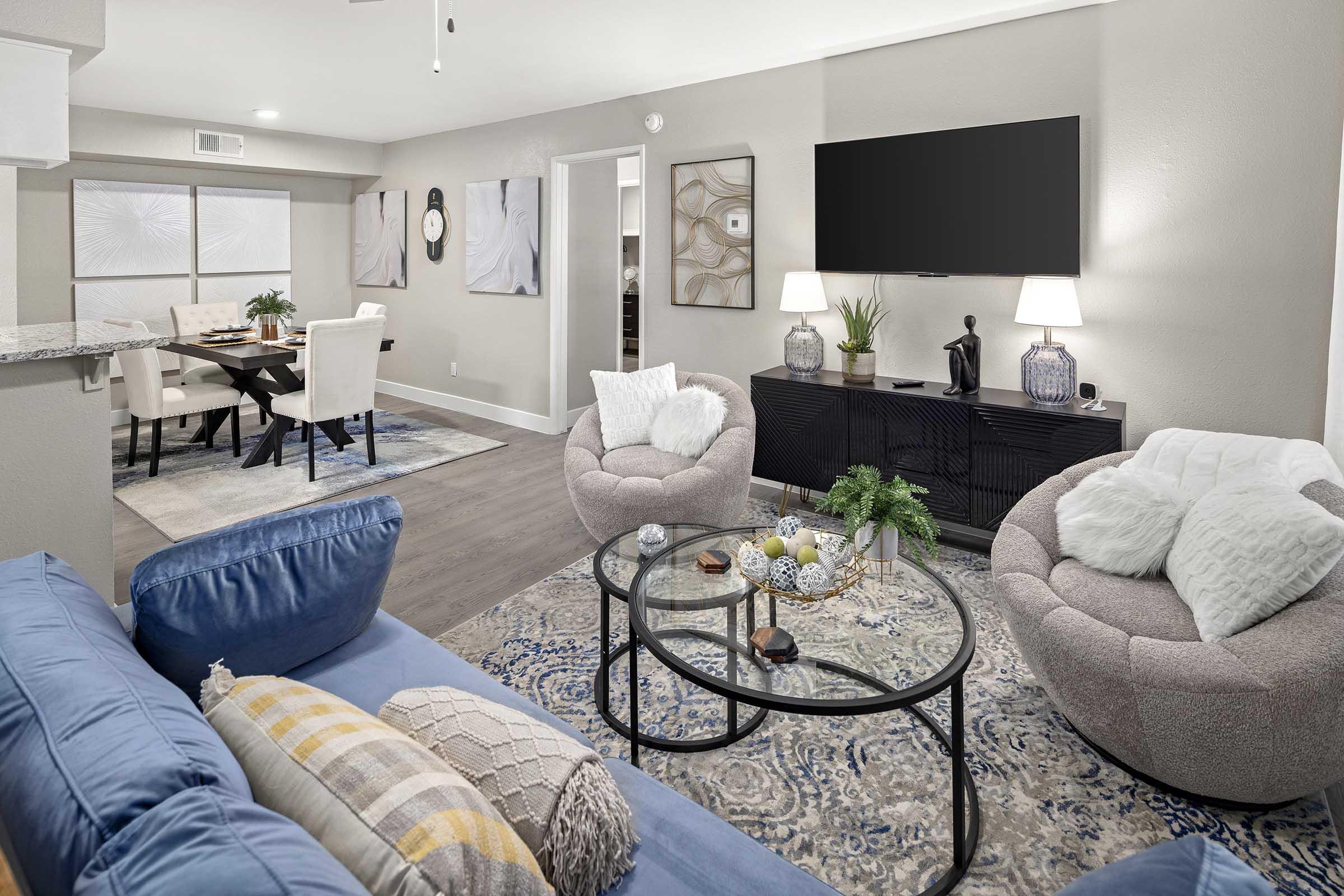 A stylish living room featuring a blue sofa with decorative pillows, a round coffee table with decorative items, and two modern chairs. The room is well-lit and includes a wall-mounted TV, a dining area in the background with a table and chairs, and artwork on the walls.