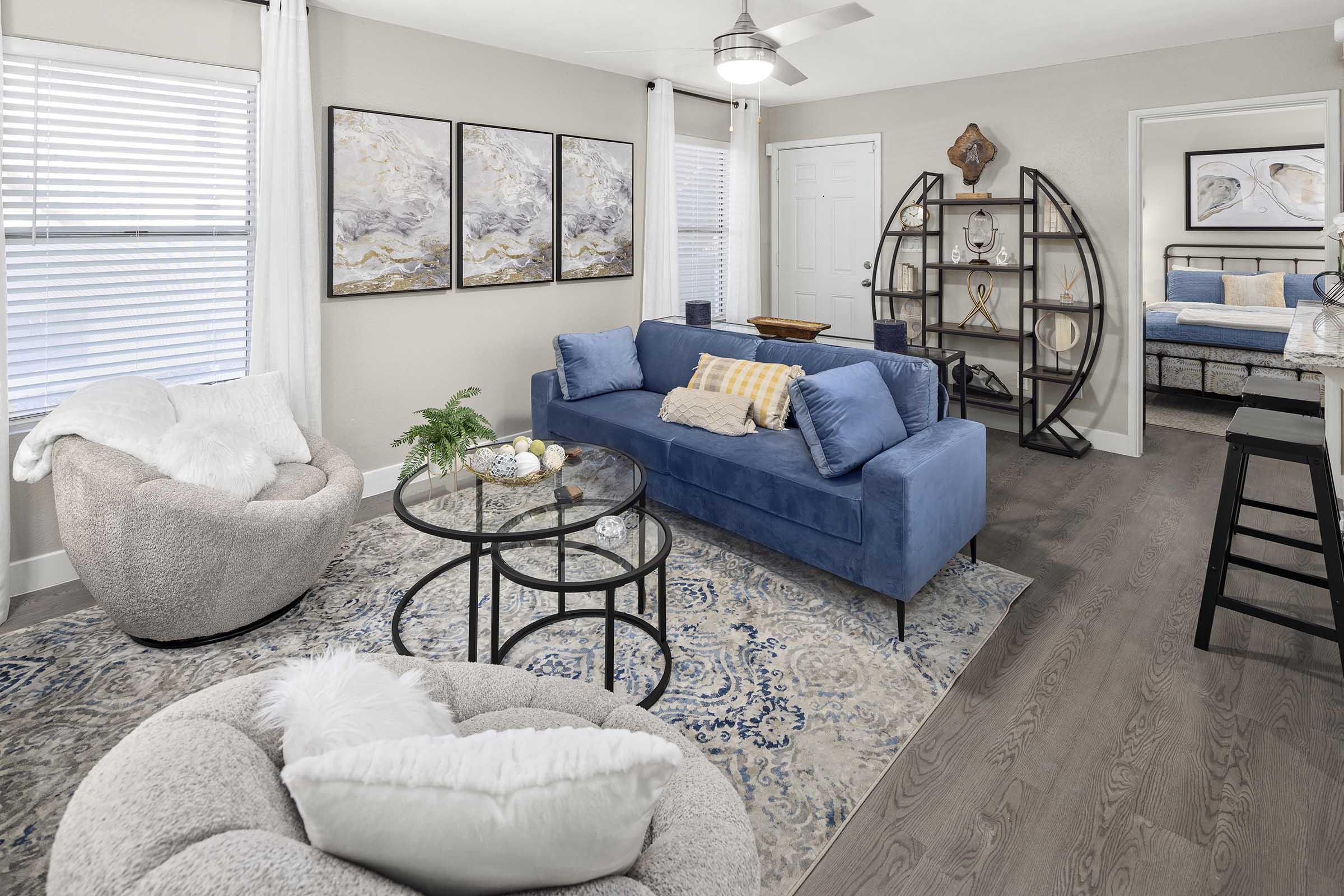 A modern living room featuring a blue sofa with decorative pillows, a round glass coffee table, and a cozy chair. The walls are adorned with abstract art, and the space includes a bookshelf and a glimpse of a bedroom in the background. Natural light filters through the windows, enhancing the inviting atmosphere.