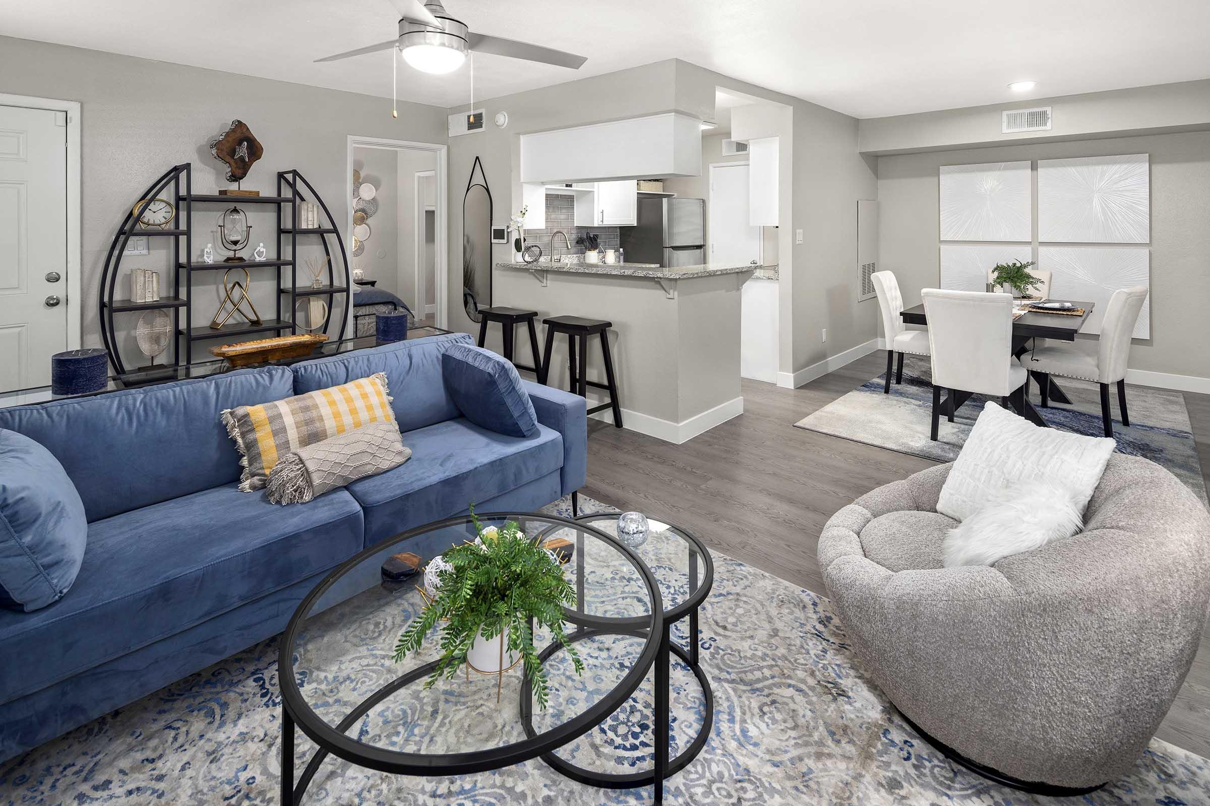 A modern living room featuring a blue sofa with patterned throw pillows, a round coffee table with a plant, and a cozy gray armchair. In the background, there's a dining area with a light-colored table and chairs, and an open kitchen visible with bar seating. The decor is contemporary and inviting.
