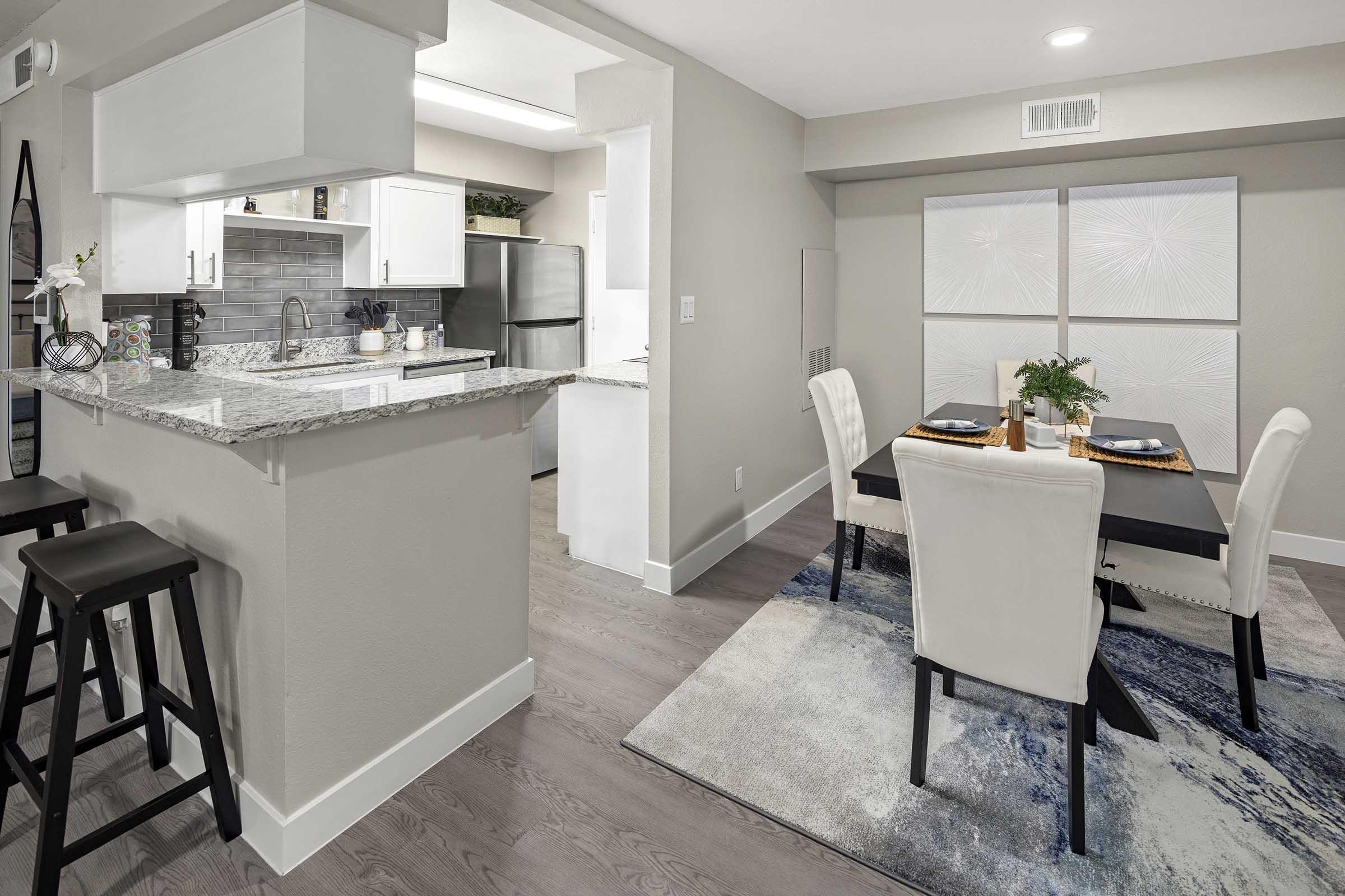 A modern kitchen and dining area featuring a granite countertop, black stools, white cabinetry, a stainless steel refrigerator, and a dining table set for four. The space has light-colored walls and a rug with a blue and white pattern, creating a stylish and inviting atmosphere.