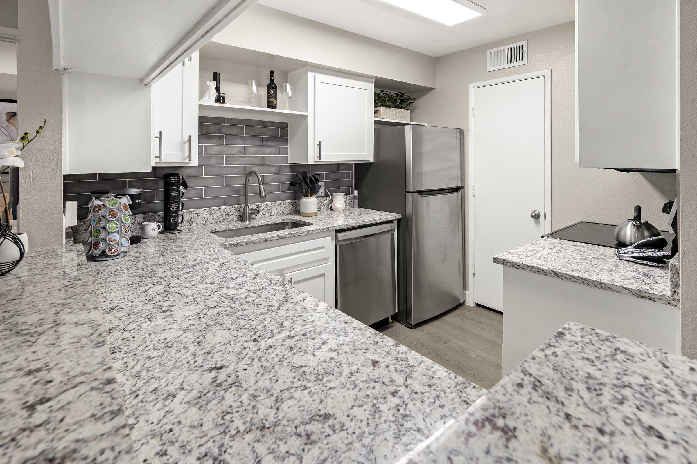 Modern kitchen featuring white cabinets, granite countertops, stainless steel appliances, and a grey-tiled backsplash. The countertop is clutter-free, with a coffee maker, decorative items, and a kettle included. Good lighting enhances the clean and contemporary design of the space.