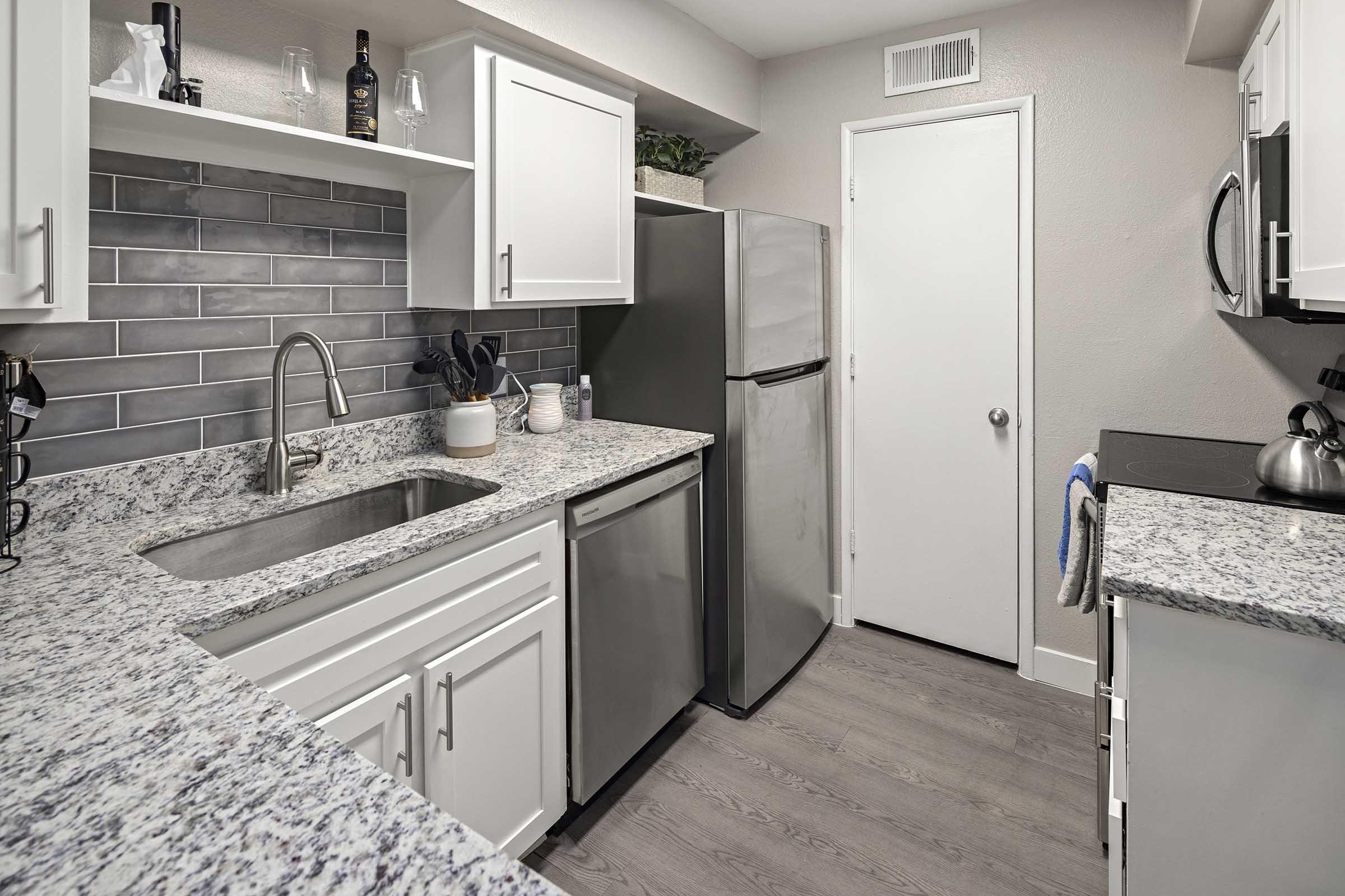 Modern kitchen featuring granite countertops, stainless steel appliances including a refrigerator and stove, white cabinetry, and gray tile backsplash. A sink is positioned under a window, and cooking utensils are neatly organized. The flooring is a light wood laminate.