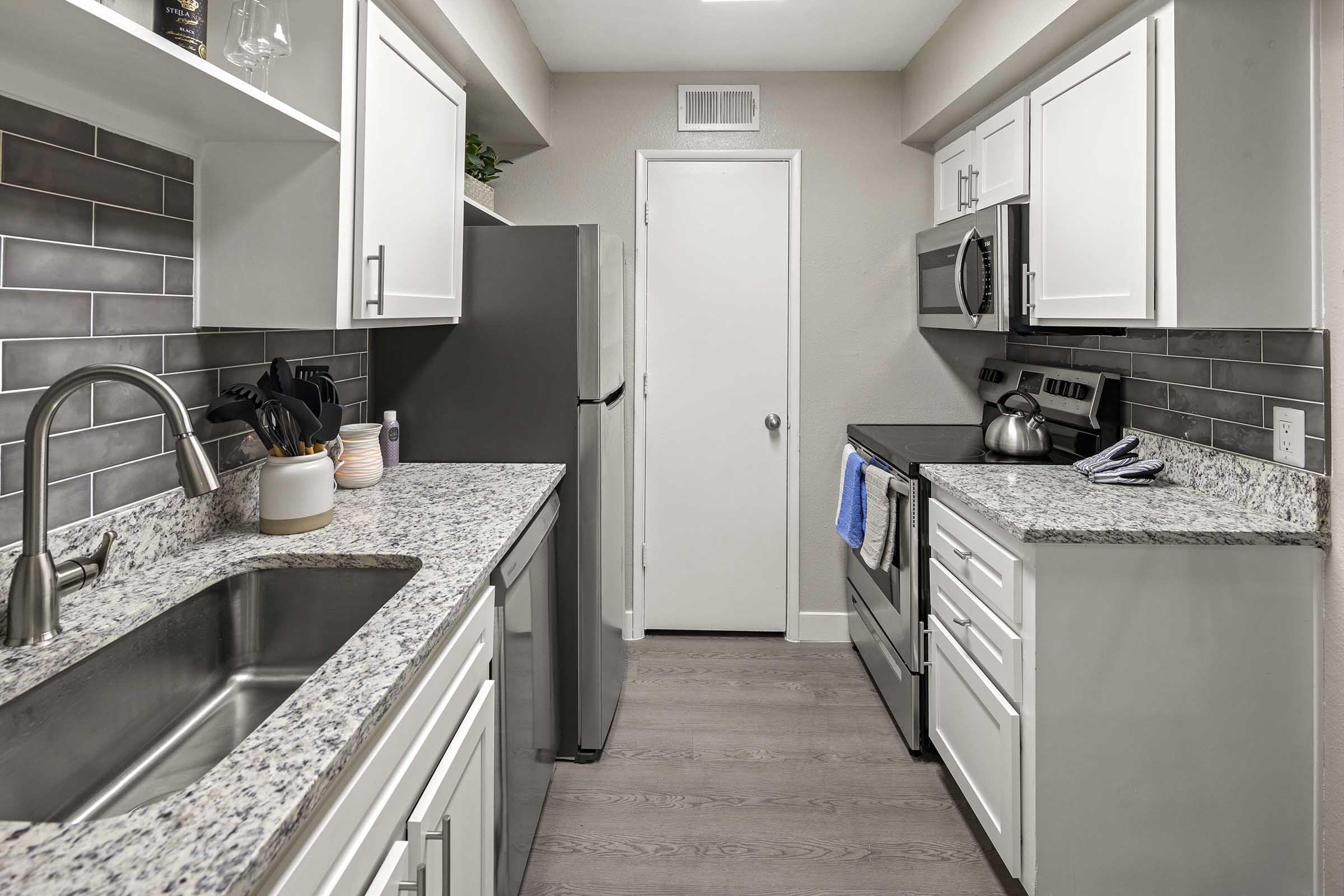 A modern kitchen featuring white cabinets, gray tile backsplash, and stainless steel appliances. The countertop is made of granite, and there is a double sink with a modern faucet. A refrigerator and an oven with a microwave are visible, along with kitchen utensils and a blue towel hanging from the oven handle.