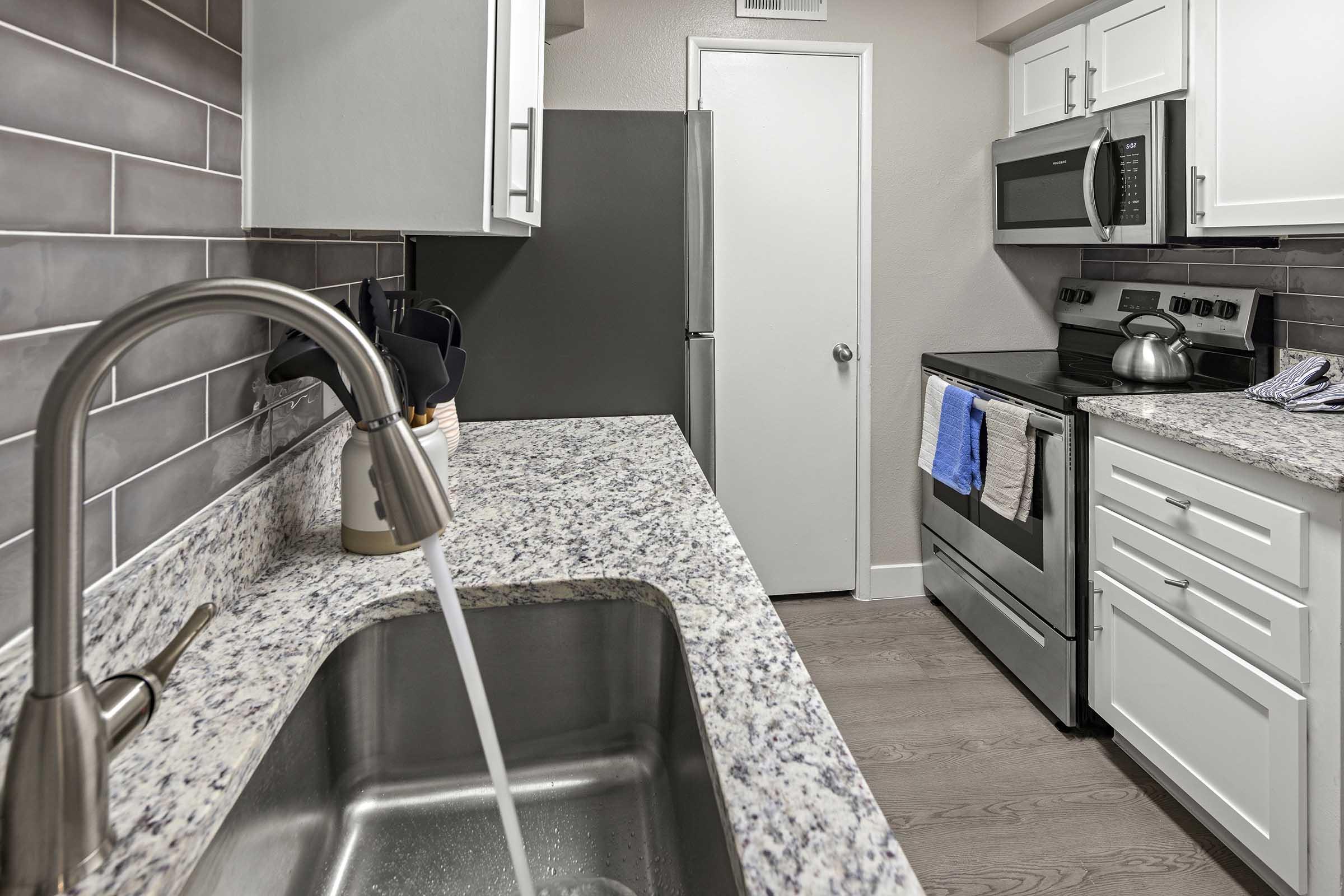 A modern kitchen featuring a granite countertop with a stainless steel sink. The sink is running with water. White cabinets and appliances, including a microwave and stove, are visible. A towel hangs on the oven, and a door leads to another room. The kitchen has gray tile backsplash and wood-style flooring.