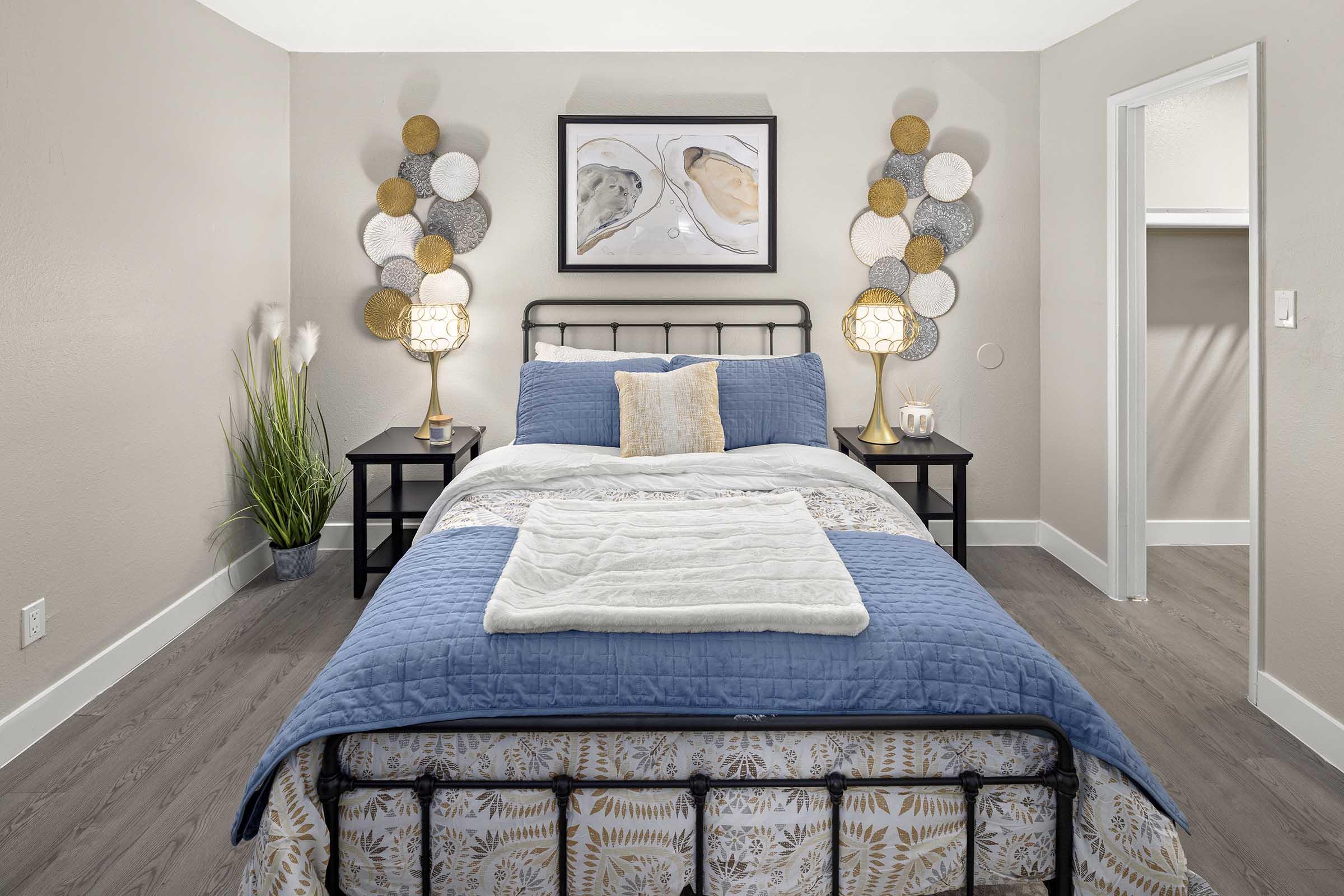A cozy bedroom featuring a metal bed frame with blue and white bedding, decorative pillows, and a plush throw blanket. There's a piece of abstract art on the wall and two lamp-lit nightstands with round wall decor. A potted plant adds a touch of greenery, and a closet is partially visible in the background.