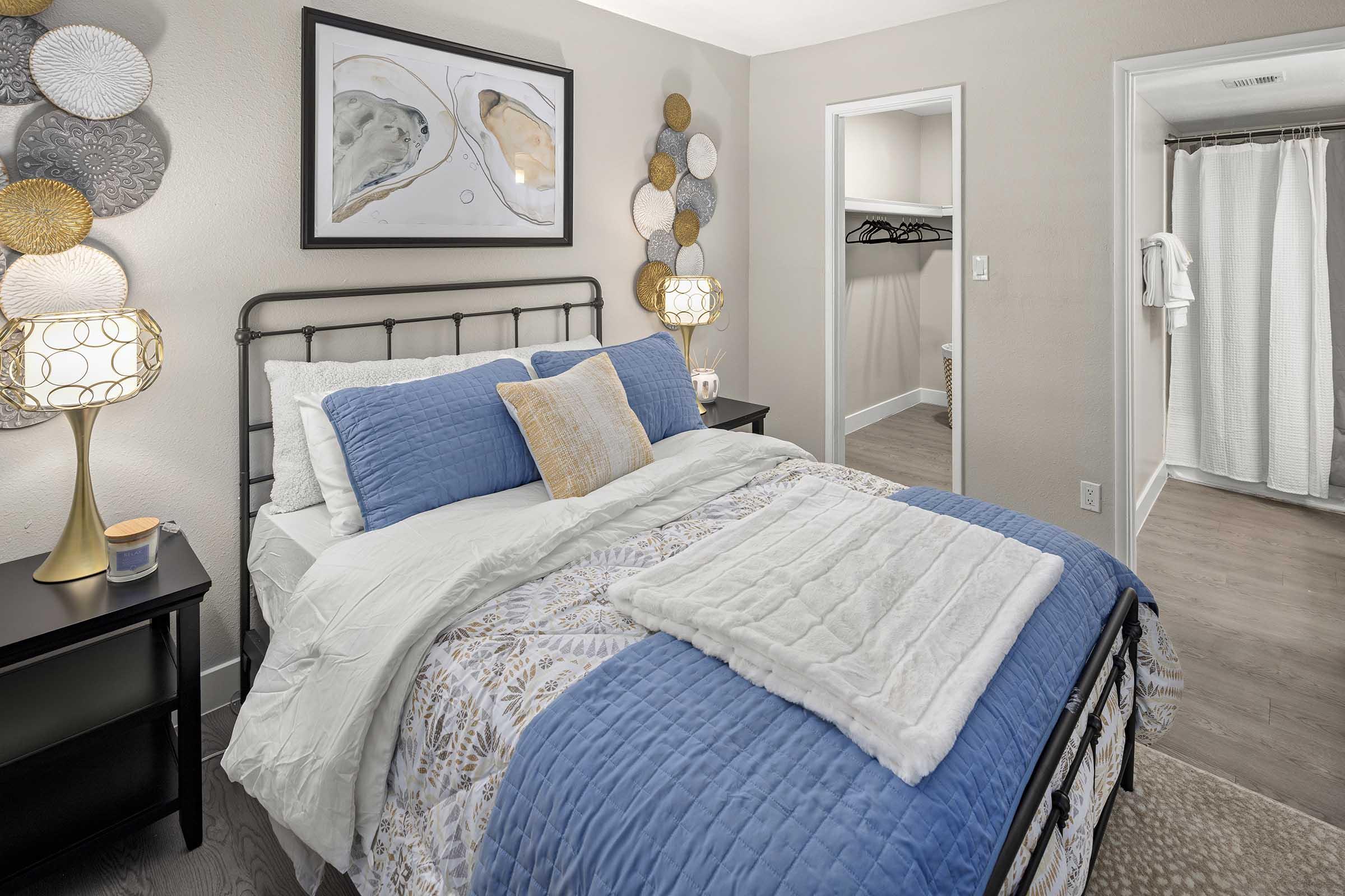 A cozy bedroom featuring a black metal bed with blue and white bedding, decorative pillows, and a fluffy blanket. There are stylish lamps on both bedside tables, wall art above the bed, and a light-colored closet space visible in the background. Soft, neutral tones enhance the inviting atmosphere.