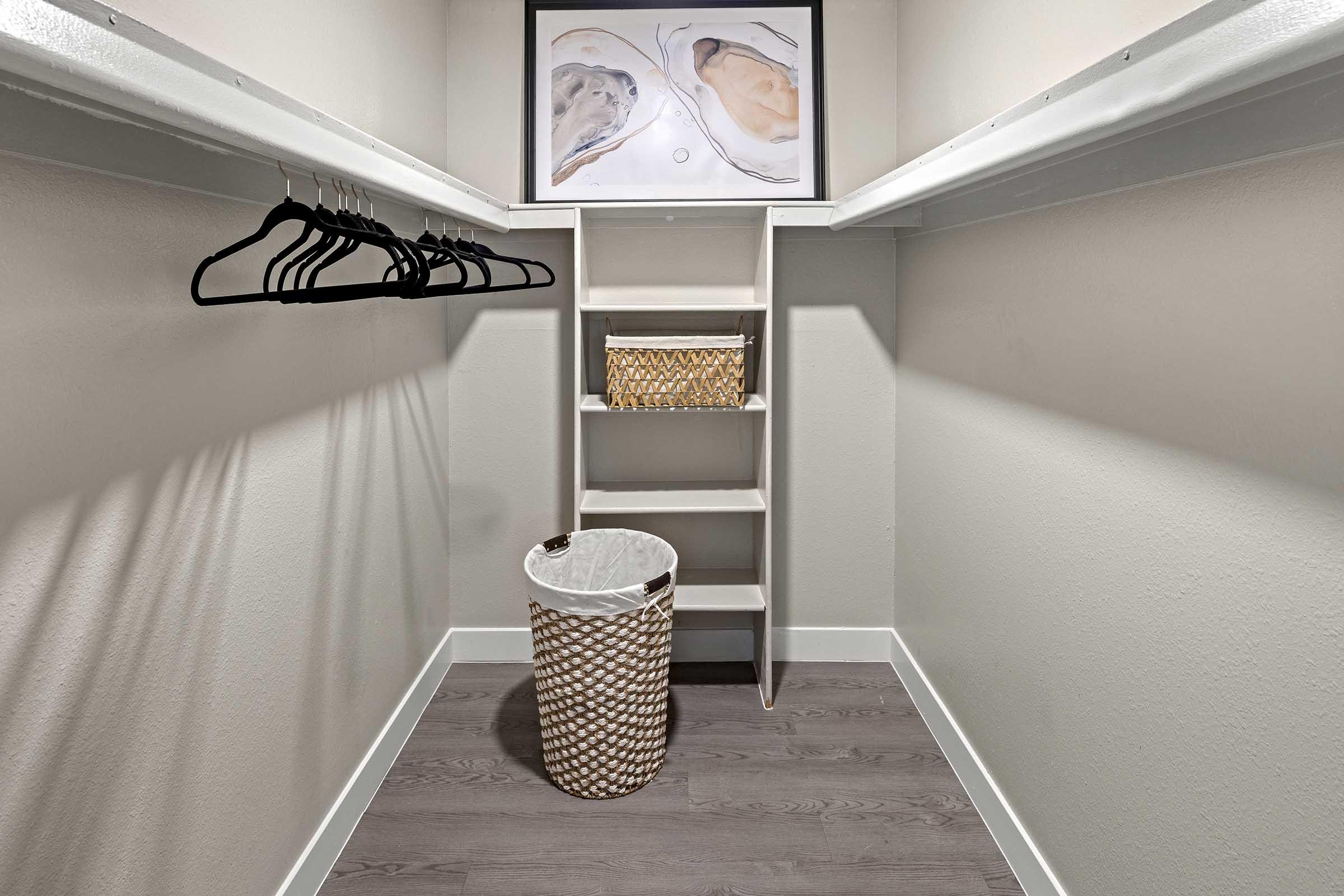 A well-organized walk-in closet featuring hangers on a rod, a small shelf unit with a decorative basket, and a laundry basket on the floor. The walls are painted in a light color, and there’s artwork displayed above the shelves, creating a clean and functional storage space.