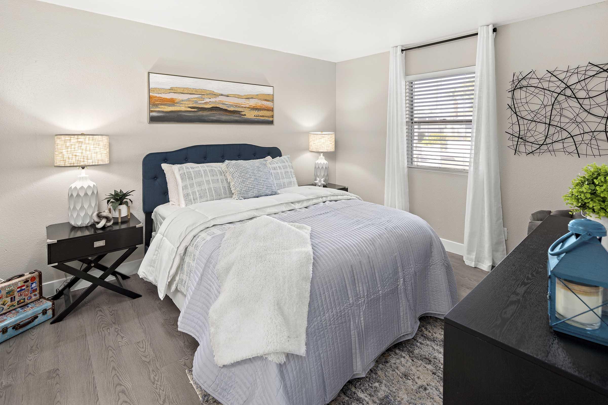 A cozy bedroom featuring a queen-sized bed with a light purple quilt and decorative pillows. Two bedside lamps provide warm lighting beside a dark nightstand. A window with white curtains allows natural light, while a wall art piece adds a modern touch. A green plant and a blue decorative lantern are on the dresser.