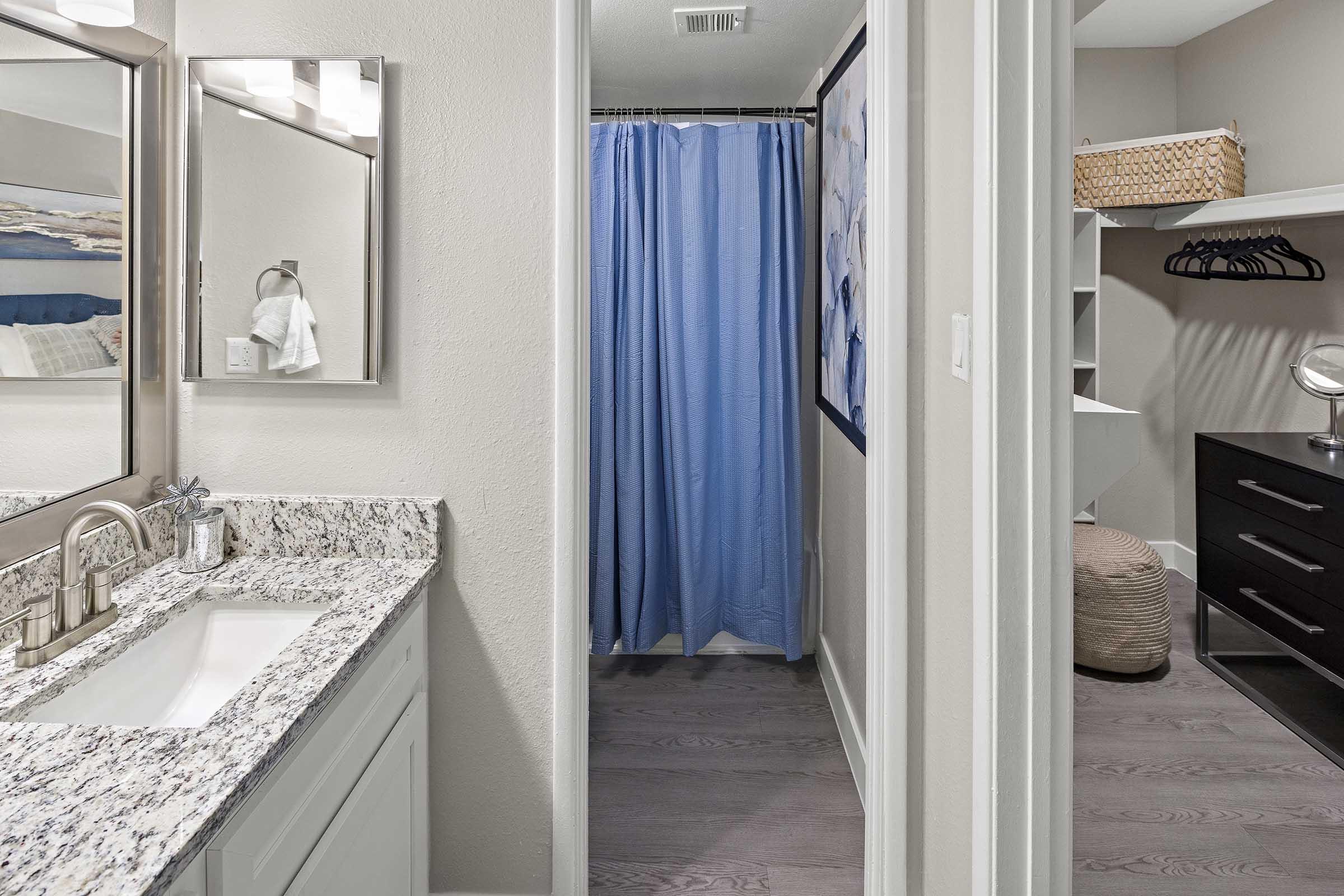 A modern bathroom featuring a granite countertop with a sink, a large mirror, and a blue shower curtain. Adjacent is a small closet area with shelving, a hanging rack, and a storage basket. The flooring is a light wood finish, and the walls are painted in neutral tones.