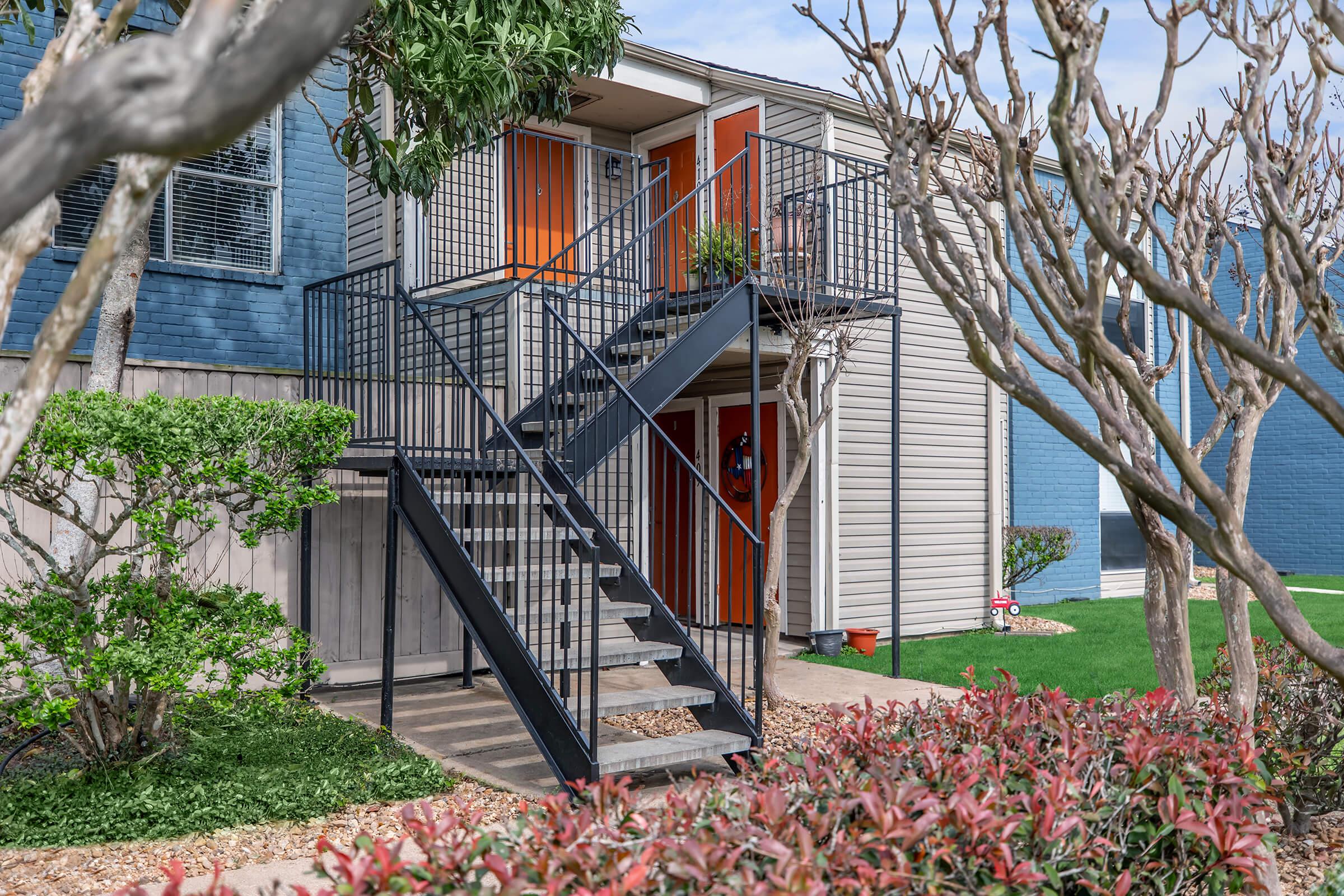 A multi-level apartment building featuring a black metal staircase leading to an upper floor. The building has blue exterior walls and orange doors, surrounded by neatly trimmed shrubs and trees. Lush green grass and decorative plants enhance the landscaping around the entrance.