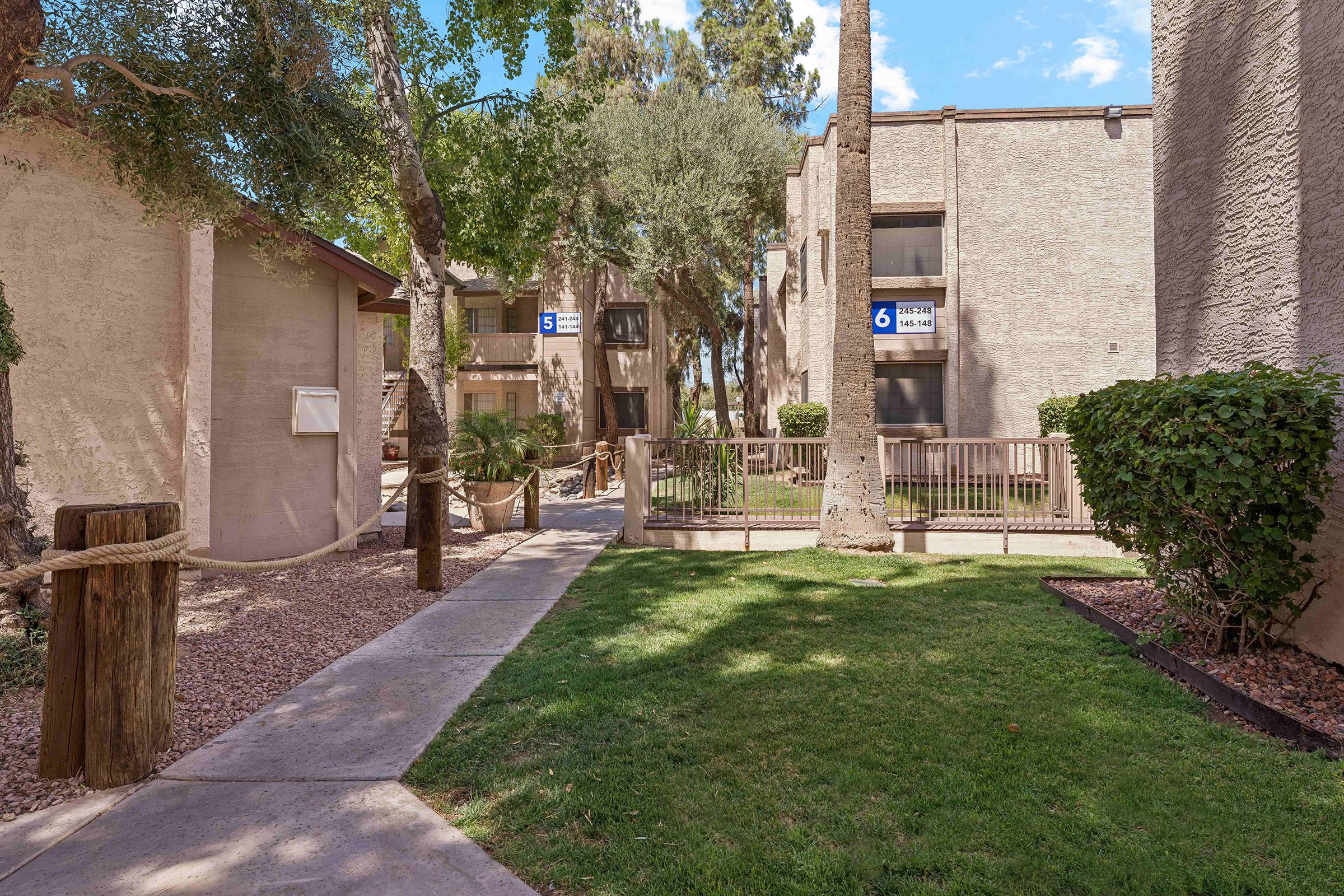 A pathway lined with greenery and trees leads through an apartment complex. Nearby buildings are partially obscured by palm trees and shrubs. A sign indicating "Unit 6" is visible. The scene is sunny with blue skies and scattered clouds, creating a pleasant outdoor atmosphere.