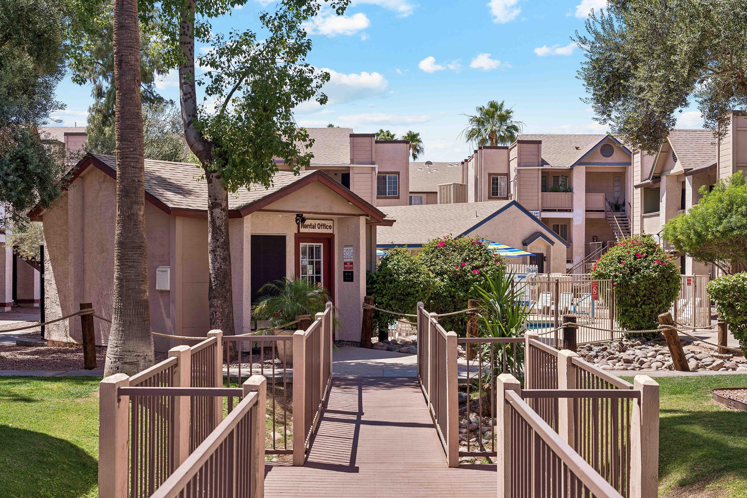 A view of an apartment complex featuring a welcoming entrance with a wooden bridge leading to the leasing office. Lush green lawns, palm trees, and flowering shrubs create a serene atmosphere, with additional buildings visible in the background. A swimming pool area is also indicated nearby. Clear blue skies are overhead.