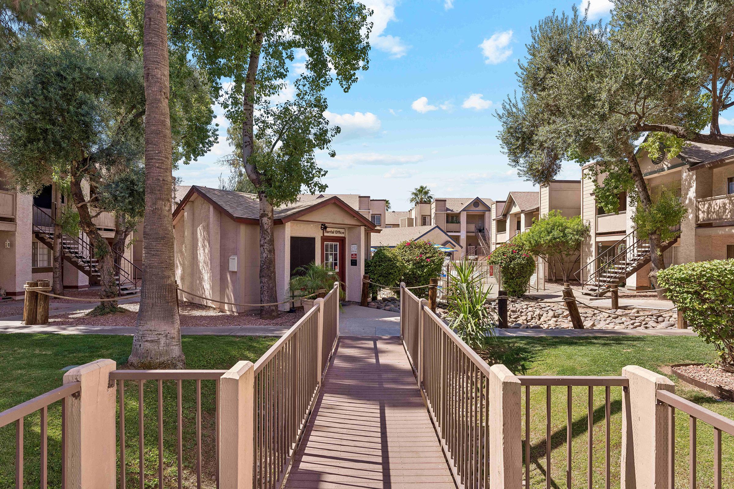 View of a residential apartment complex featuring a wooden walkway leading to an entrance. The area is surrounded by lush greenery, including palm trees and shrubs, with buildings on either side and a clear blue sky overhead. The setting appears inviting and well-maintained.