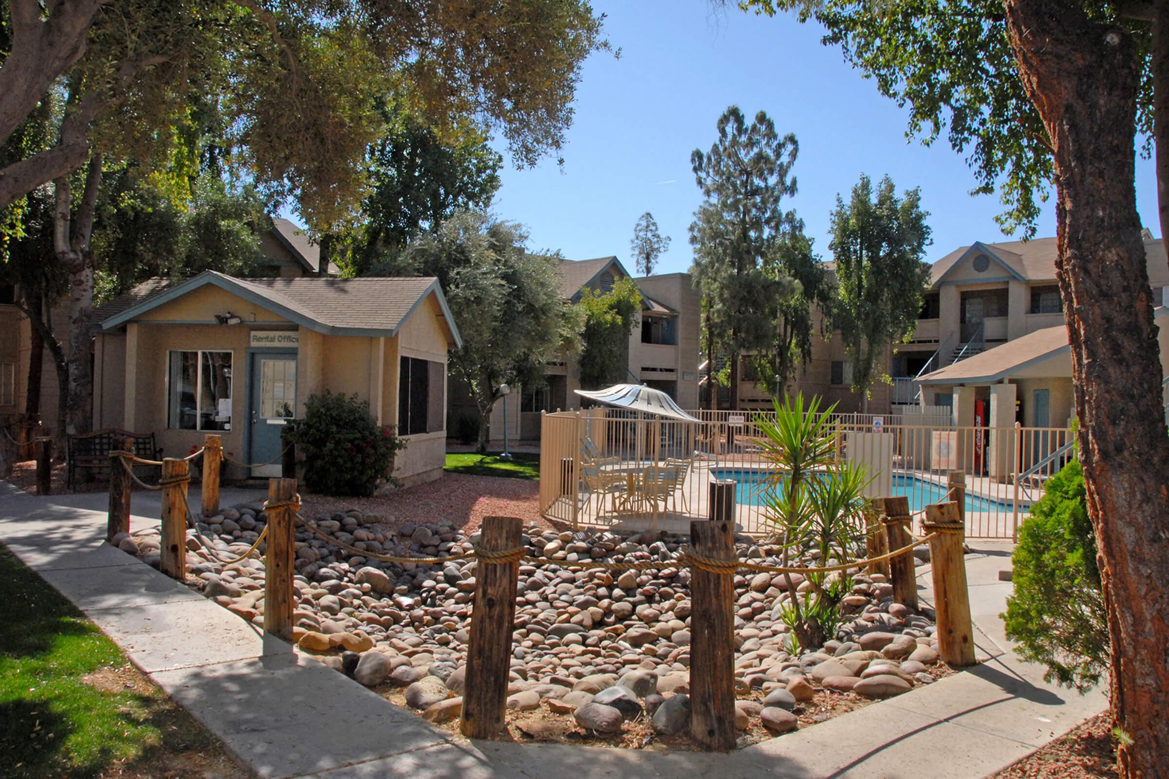 A tranquil residential area featuring a small swimming pool surrounded by a fence, a seating area with palm plants, and landscaped gardens with rocks.
