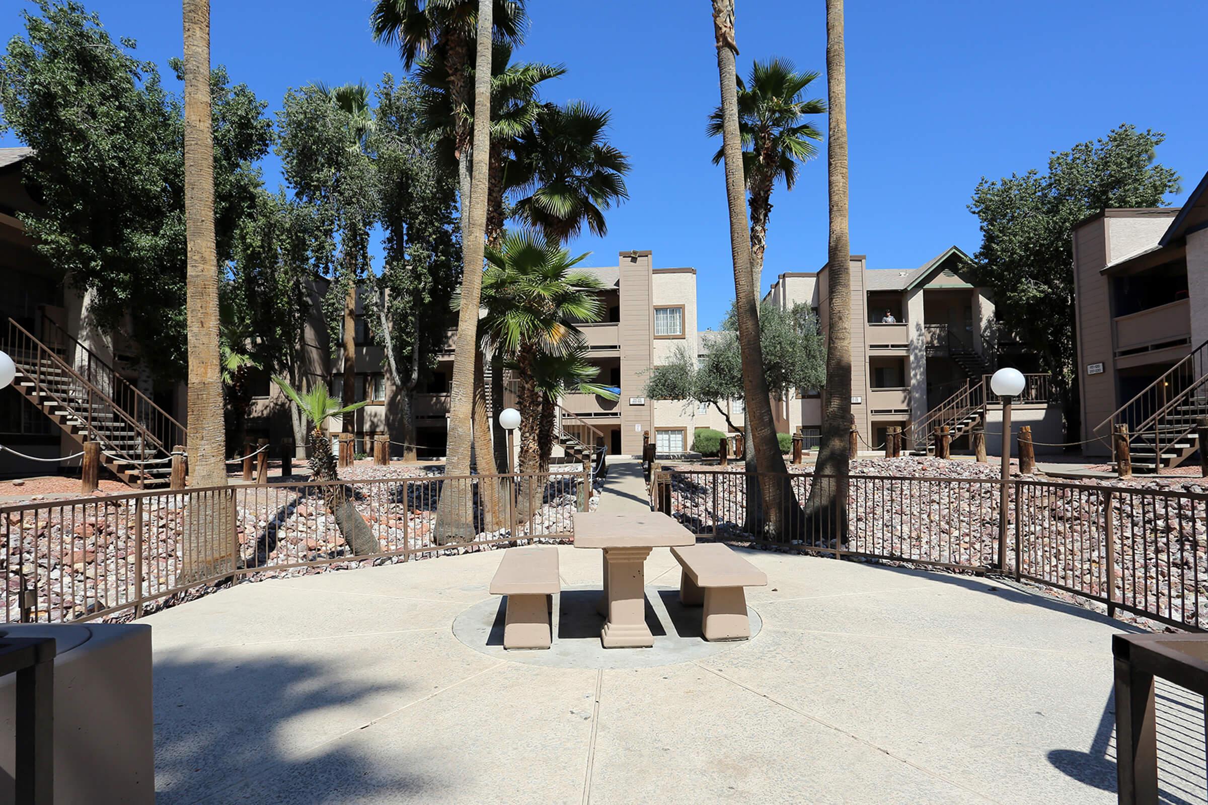 A courtyard area surrounded by residential buildings, featuring palm trees and a circular stone table with benches.