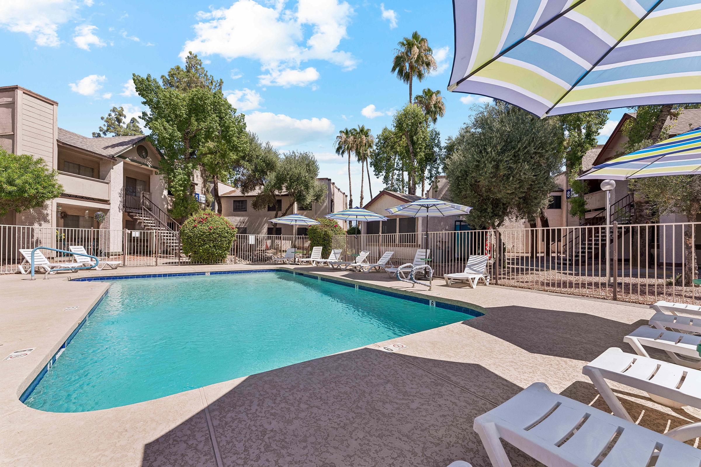 A serene pool area surrounded by lounge chairs and umbrellas. Lush green trees and palms provide shade in the background, with nearby apartment buildings visible. The clear blue water reflects a bright sky with fluffy clouds, creating a relaxing atmosphere.