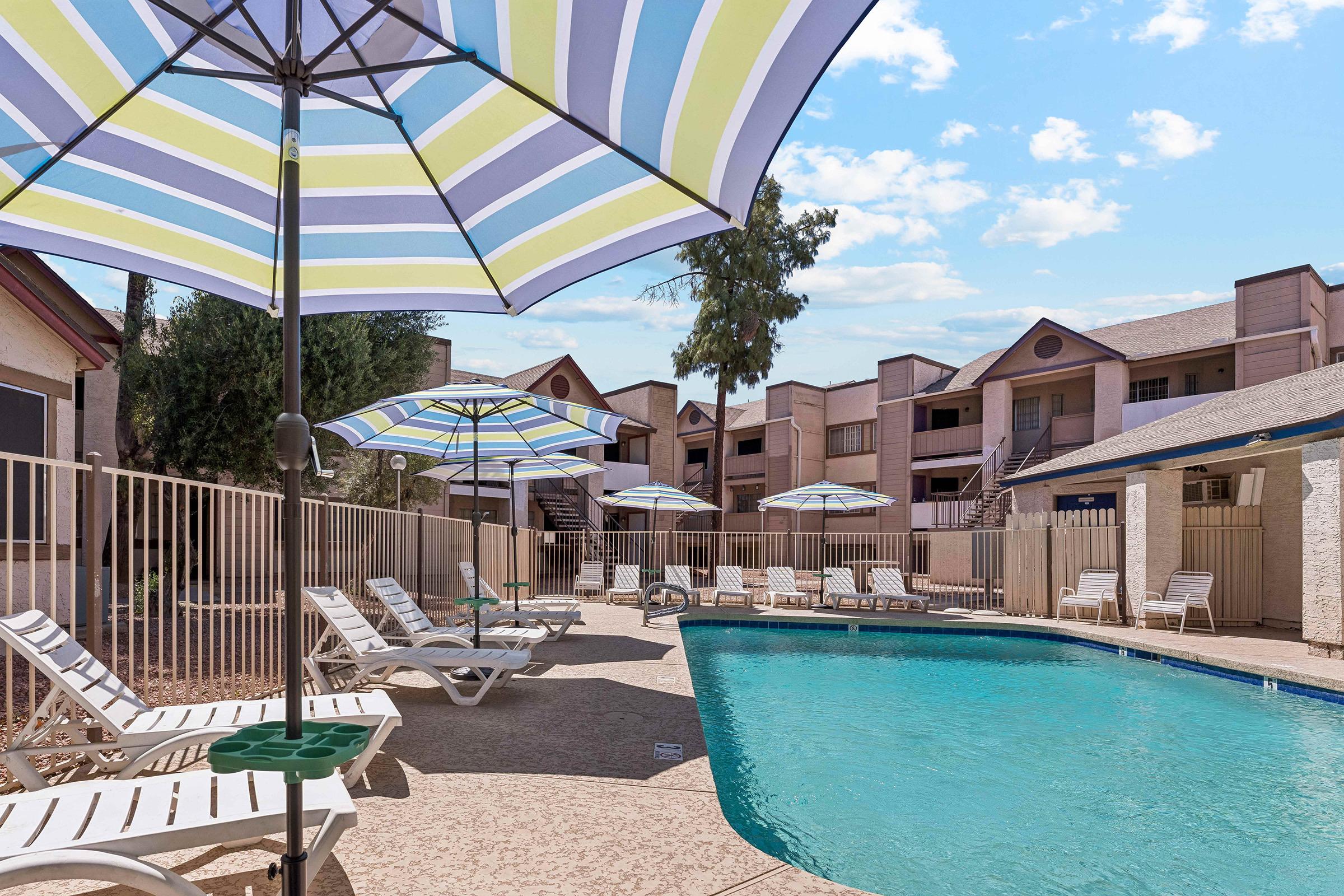 An inviting swimming pool surrounded by lounge chairs and colorful umbrellas, set in a well-maintained outdoor area. Nearby apartments are visible in the background, with clear skies above, creating a relaxing atmosphere for leisure and recreation.