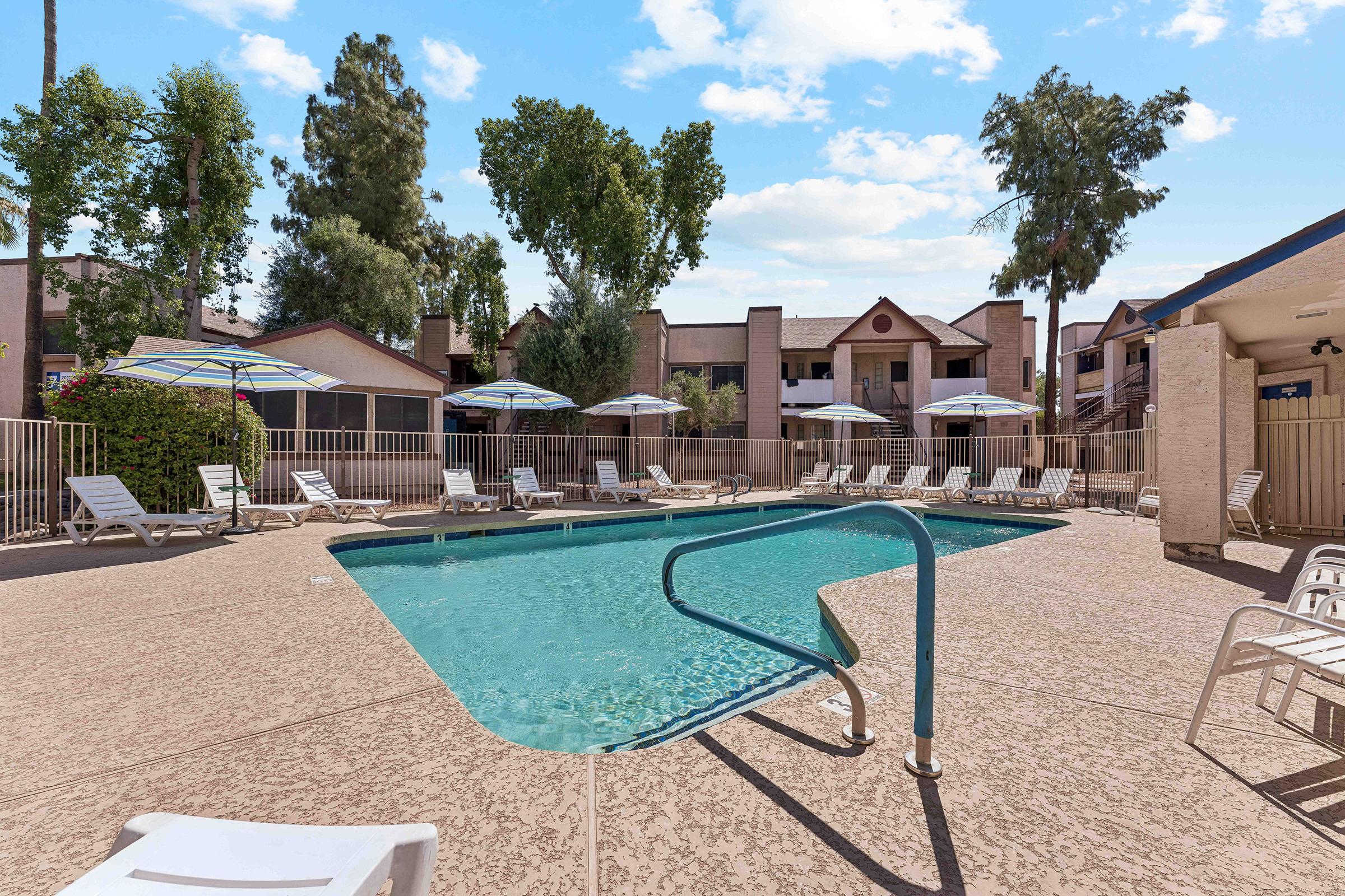 A bright, sunny pool area featuring a clean, blue swimming pool surrounded by lounge chairs and colorful umbrellas. Lush green trees are visible in the background, and there are several apartment buildings nearby. The scene conveys a relaxed and inviting atmosphere.