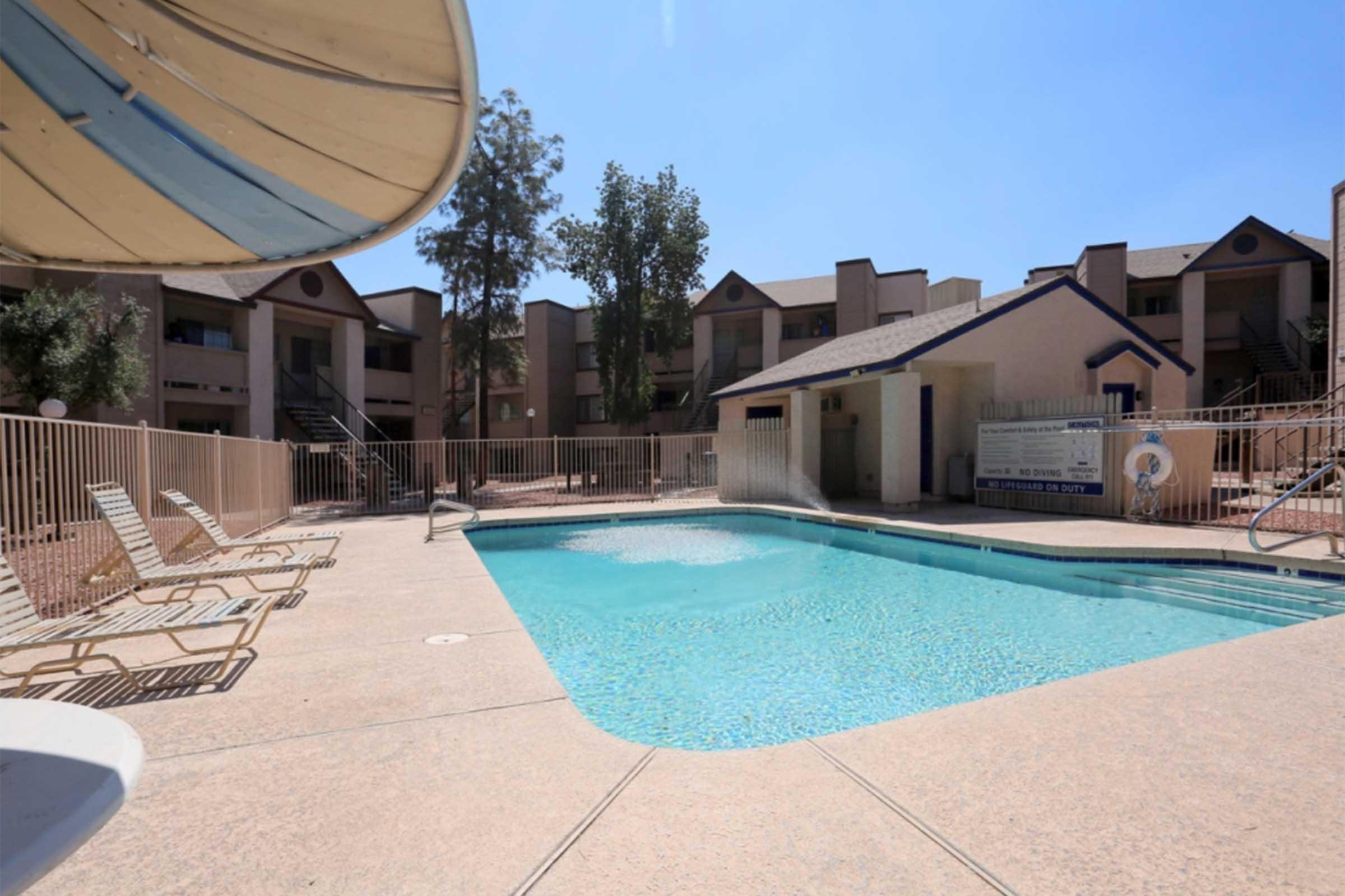 A clear blue swimming pool surrounded by lounge chairs and a fenced area, with several apartment buildings in the background.