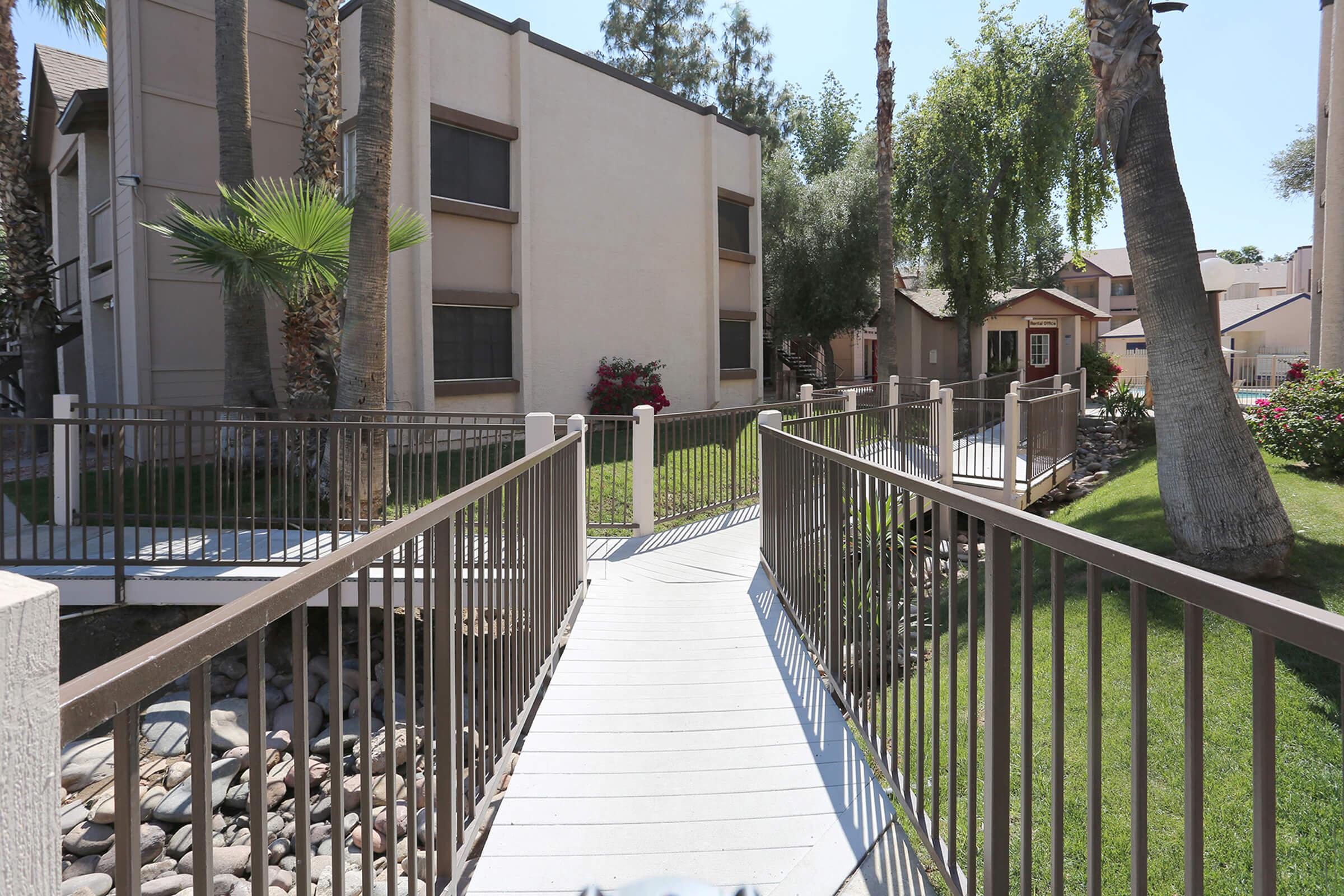 A view of a residential complex featuring a walkway bridge surrounded by greenery, palm trees, and flower beds.
