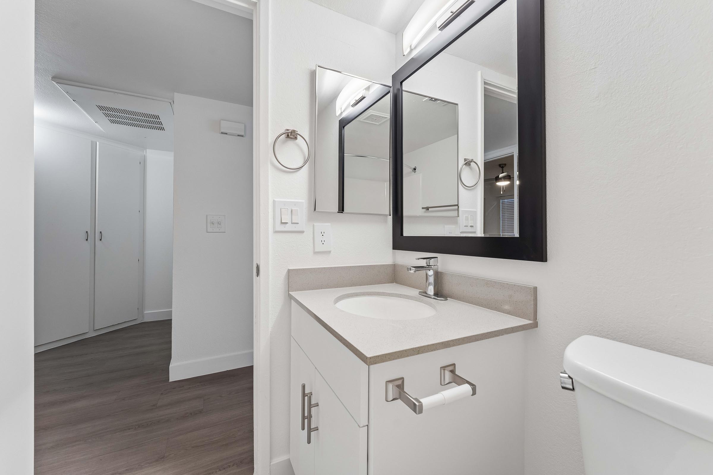 Modern bathroom featuring a sleek vanity with a sink, a wall-mounted mirror, and a chrome towel ring. The walls are painted white, and the flooring is a wood-like laminate. A toilet is visible on the right, while a doorway leads to a well-lit room in the background.