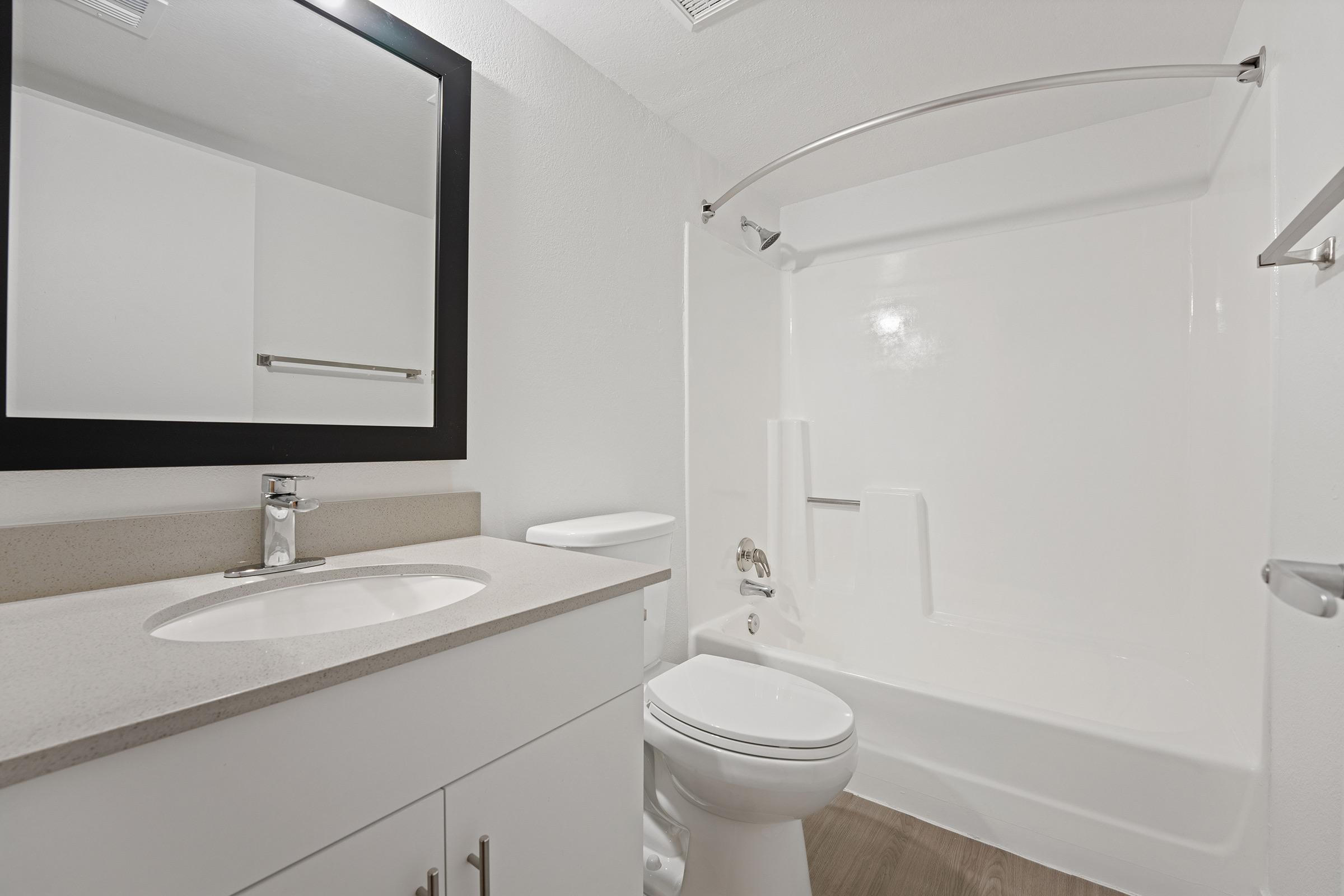 A modern bathroom featuring a clean, white design. It includes a sink with a rectangular mirror above, a toilet, and a shower area with a bathtub. The countertop is light-colored, and the flooring is a wood-like material, creating a bright and airy atmosphere.