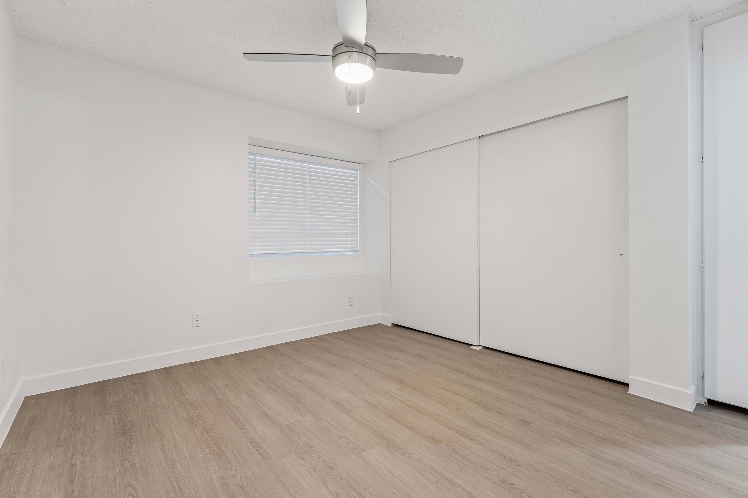 A spacious, empty room with light-colored walls and a wood-like floor. It features a window with blinds, a ceiling fan, and sliding closet doors. The overall atmosphere is bright and modern, ideal for a fresh start or new decor.