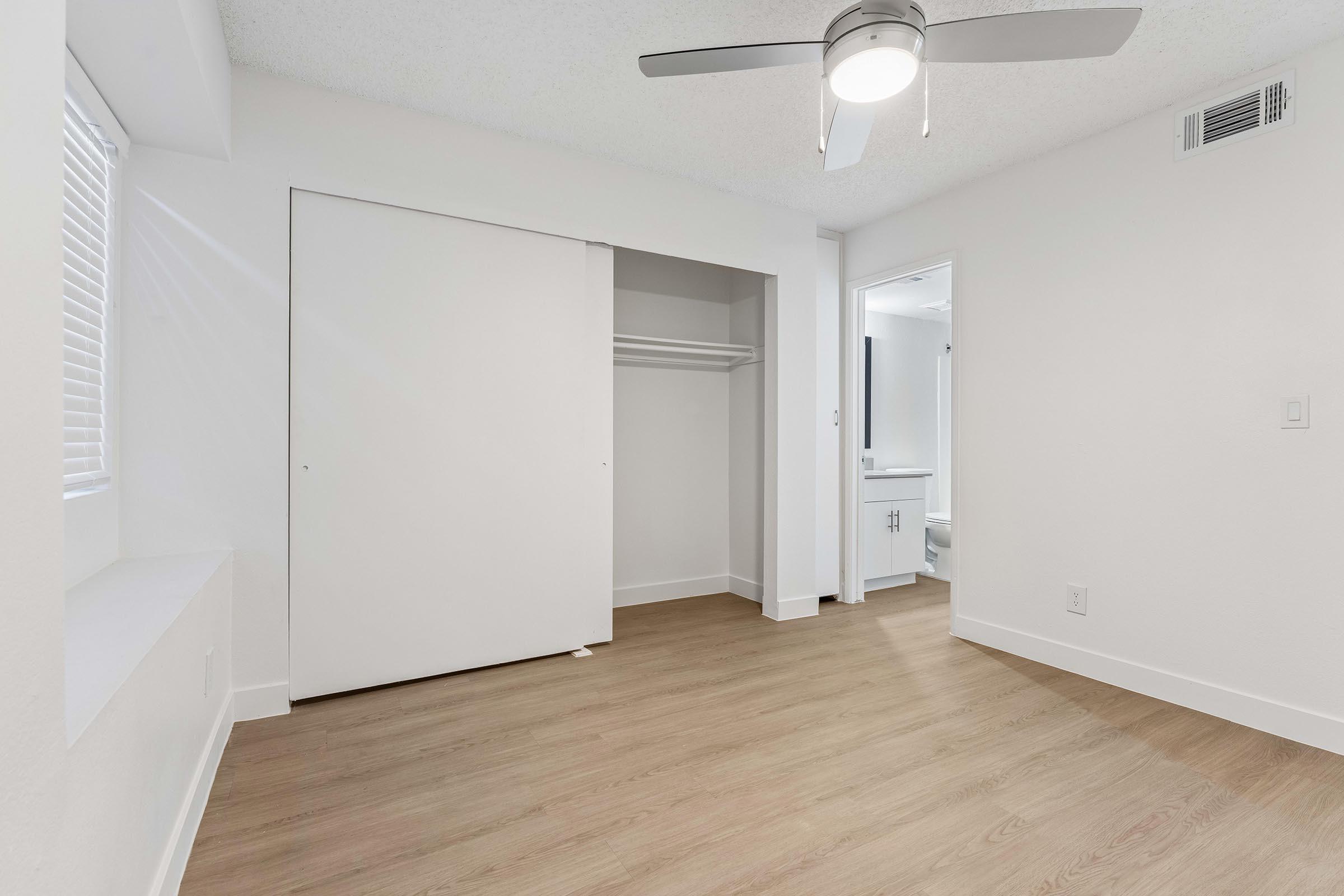 A spacious, empty room featuring light-colored walls and a wood-like floor. There is a ceiling fan, a large closet with sliding doors, and a small window with blinds. A doorway is visible on the right, leading to a bathroom area, which includes a glimpse of white fixtures. Natural light fills the space.