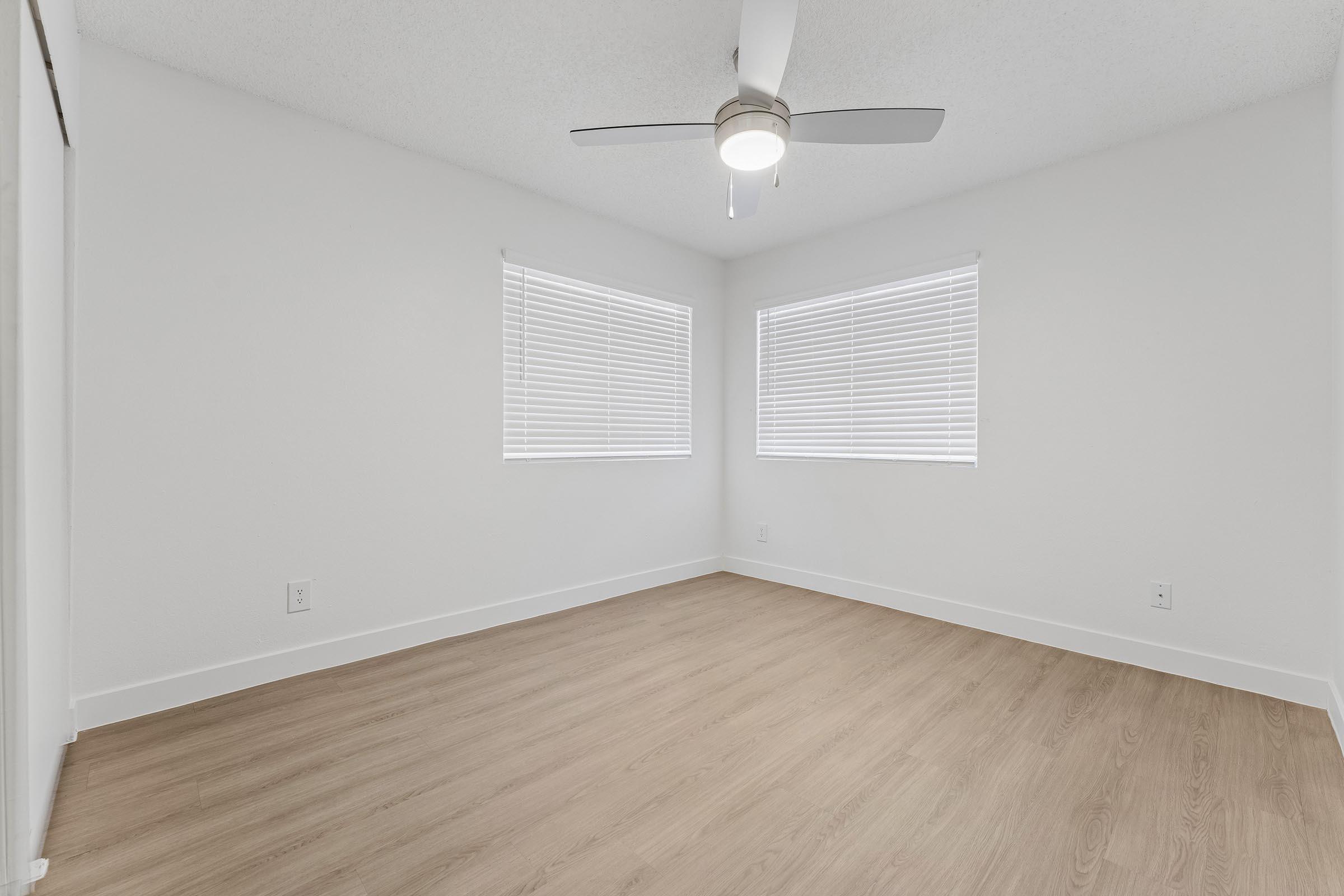 A bright, empty room with light-colored walls and wood-like flooring. It features two windows with blinds allowing natural light in and a modern ceiling fan. The space is clean and minimalistic, ideal for various uses such as a bedroom or office.