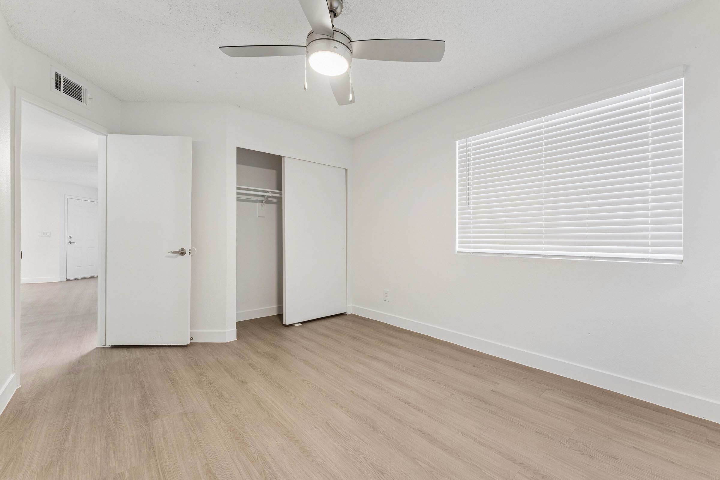 A well-lit, empty room featuring light wood flooring, a ceiling fan, and a window with white blinds. There is a closet with a sliding door on one side and an open door leading to another area of the house. The walls are painted white, creating a clean and modern look.