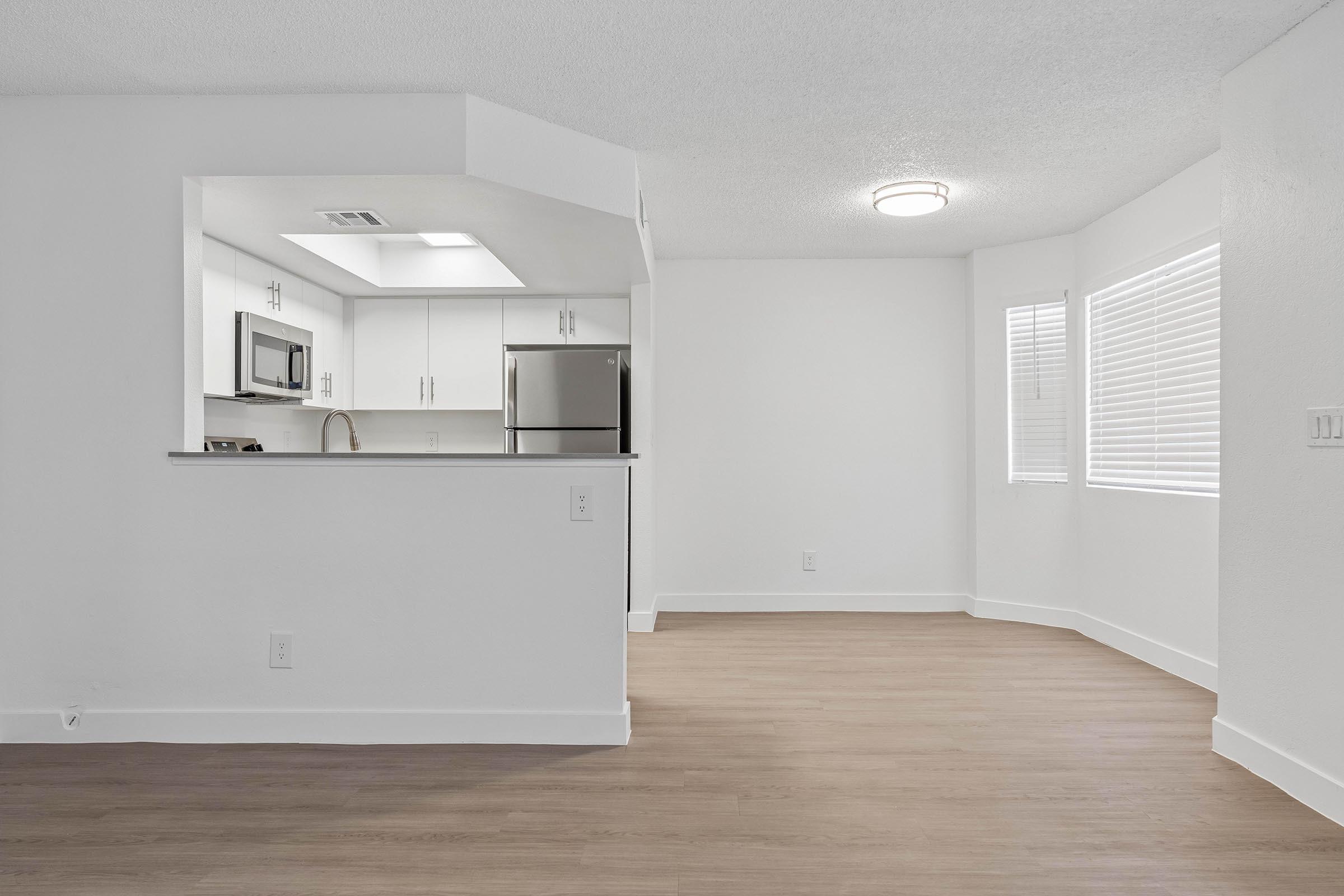 A well-lit, modern kitchen and living area featuring white walls, light wood flooring, and large windows. The kitchen includes a refrigerator, microwave, and open counter space, creating a spacious and airy feel. The area is uncluttered, with natural light enhancing the inviting atmosphere.