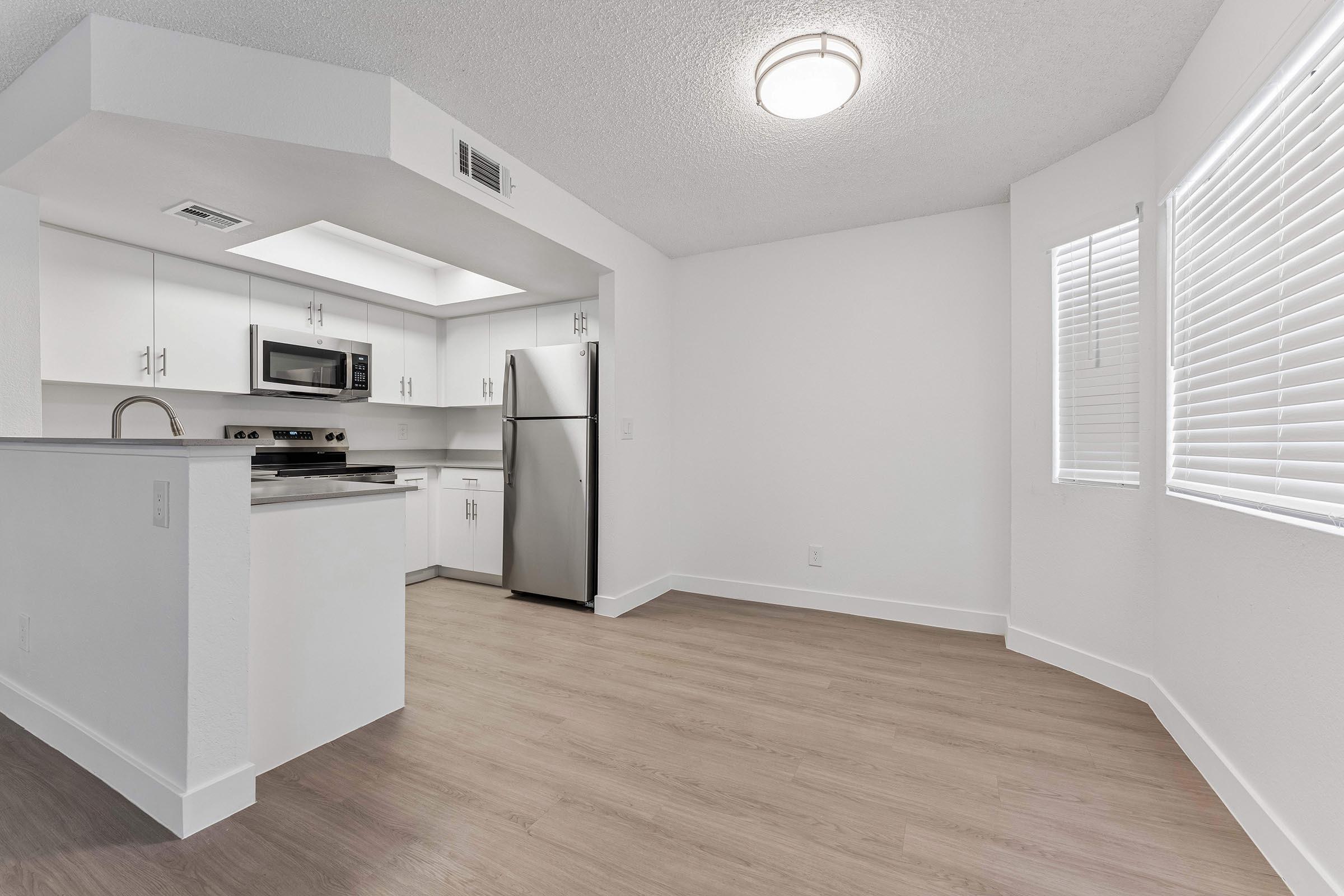 Modern kitchen with white cabinetry, stainless steel appliances including a microwave and refrigerator, and a light-colored floor. The space is bright with natural light coming through the window, and features a light fixture on the ceiling. It has an open layout, creating a spacious and inviting atmosphere.