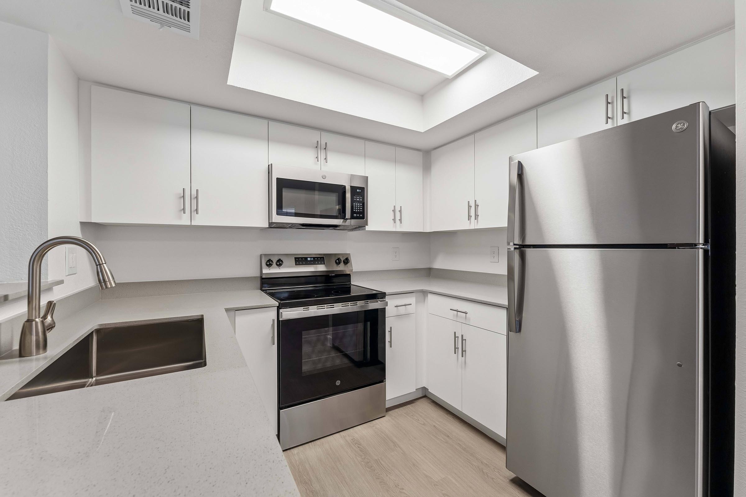 Modern kitchen featuring white cabinetry, stainless steel appliances including a microwave, oven, and refrigerator, and a sleek sink. A skylight provides natural light, and the countertops are light-colored, enhancing the spacious feel of the room. The flooring is designed for a contemporary look.