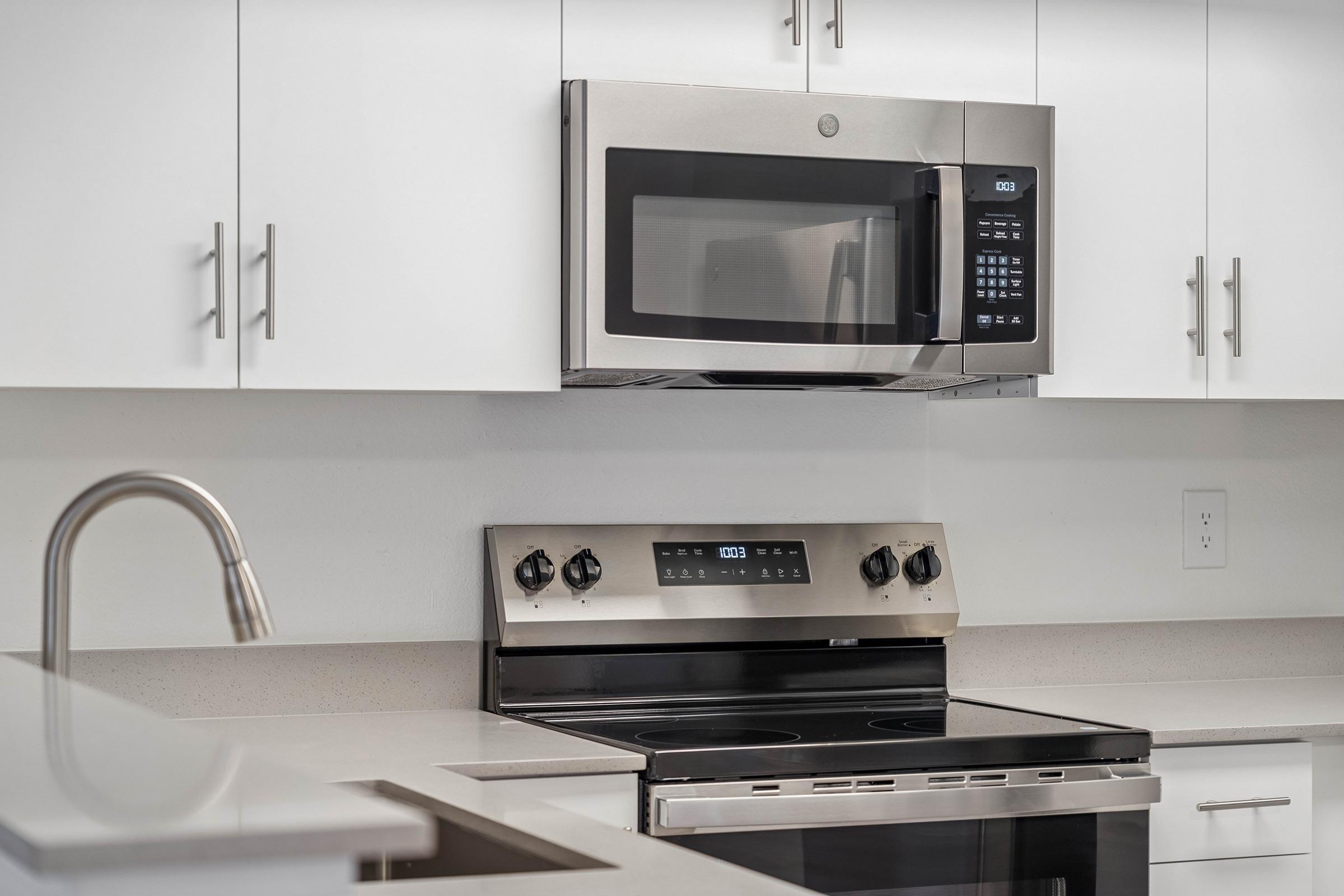 A modern kitchen featuring white cabinets, a stainless steel microwave mounted above an electric stove. The stove has a sleek, dark glass top and an oven below, set against a clean, minimalistic countertop. A polished stainless steel sink is partially visible in the foreground.