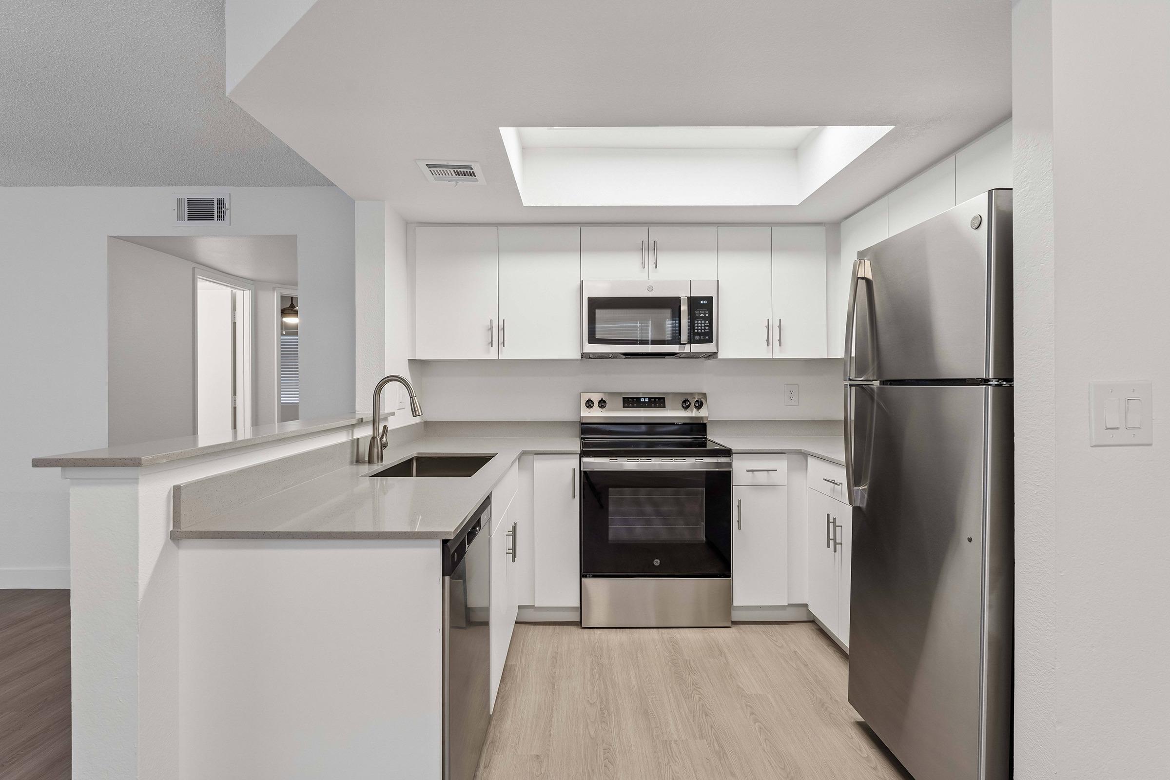 Modern kitchen featuring white cabinetry, stainless steel appliances including a microwave, oven, and refrigerator. The countertops are light-colored, with a spacious layout and natural light coming from a skylight. The flooring is a light wood finish, creating a bright and inviting atmosphere.