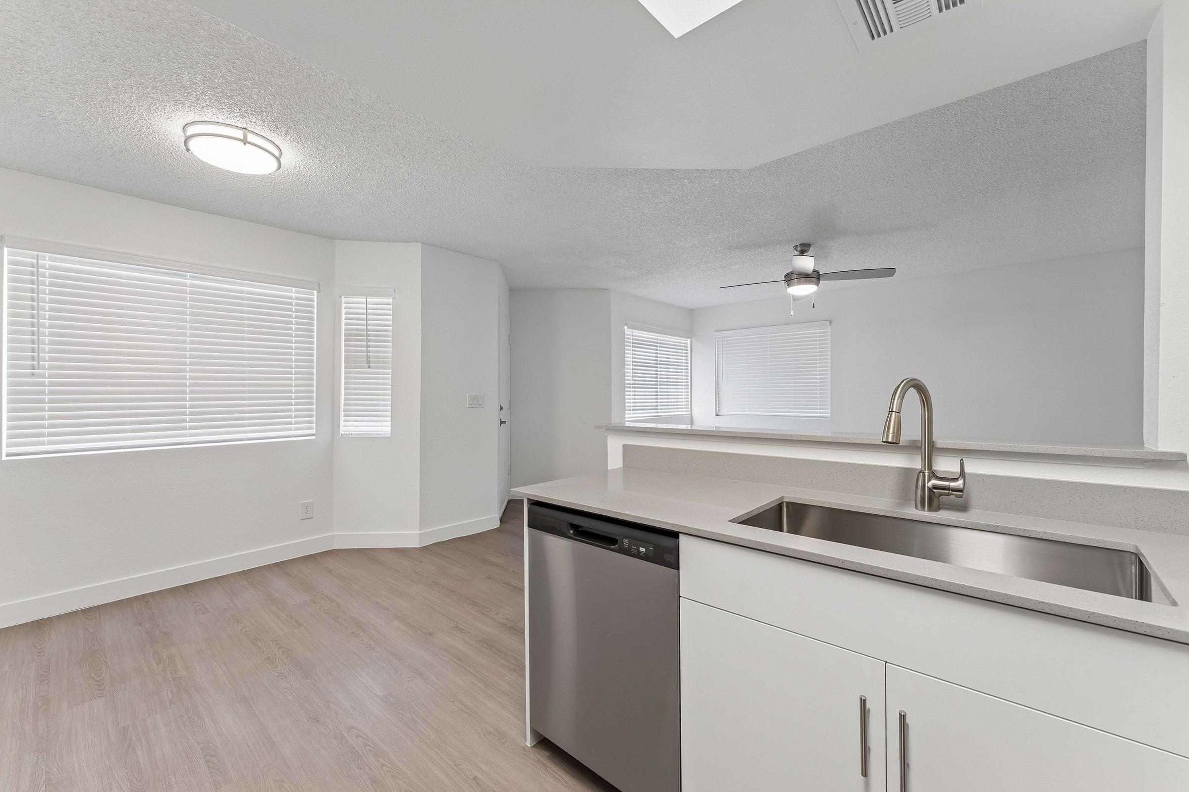 Modern kitchen and living area featuring a stainless steel sink and dishwasher, light-colored cabinets, and large windows with blinds allowing natural light. The ceiling has a light fixture and a ceiling fan, with a neutral-colored textured ceiling and wood-like flooring.
