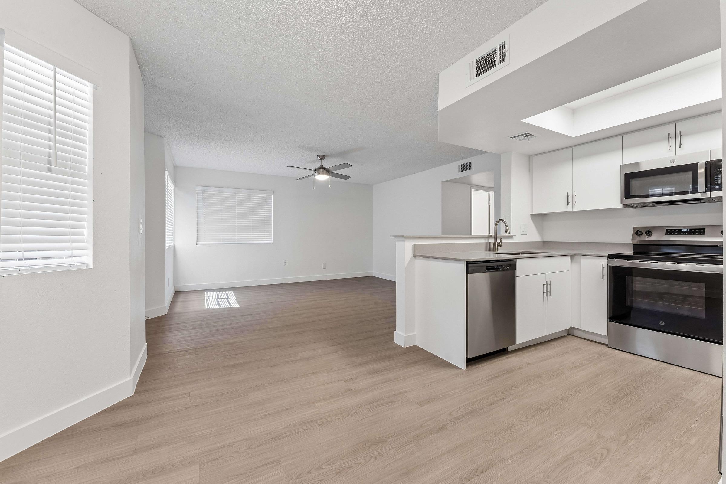Modern open-concept living space featuring a kitchen with stainless steel appliances, including a dishwasher and oven. The area is well-lit with natural light from the windows, has light-colored walls, and a ceiling fan. The flooring is a light wood laminate, creating a clean and inviting atmosphere.