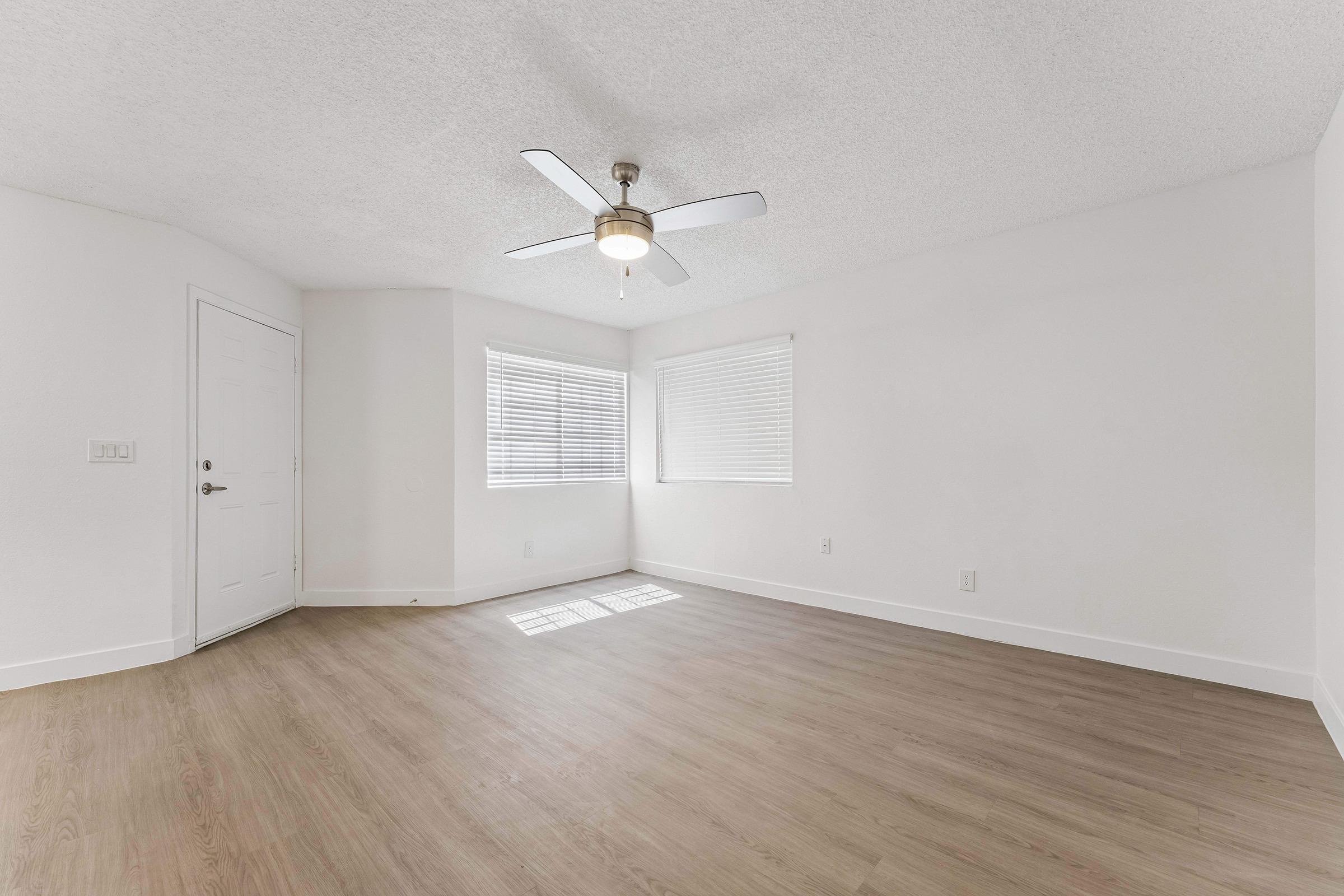 A spacious, well-lit room with light wood flooring, a ceiling fan, and large windows with white blinds. The walls are painted white, and there is a door on the left side leading outside. The room has a clean, modern appearance, perfect for various uses.