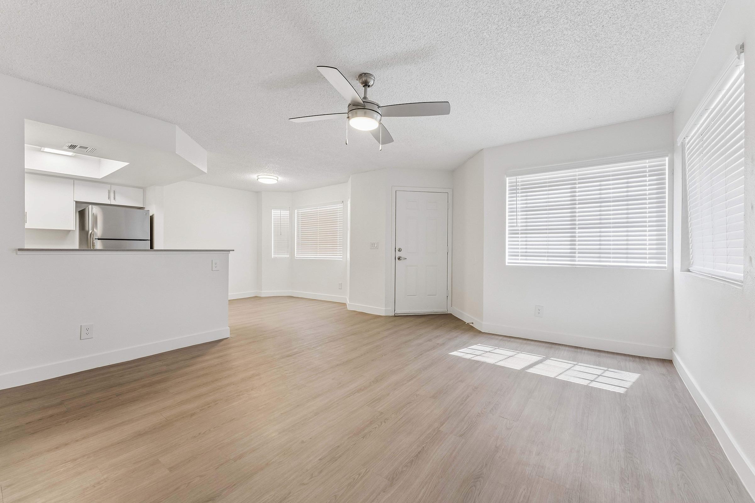 Bright, airy interior of a modern apartment featuring light-colored walls, a ceiling fan, and large windows with blinds. The open layout includes a kitchen area visible to the left and a welcoming entry door. The flooring is a light wood finish, creating a warm and inviting atmosphere.