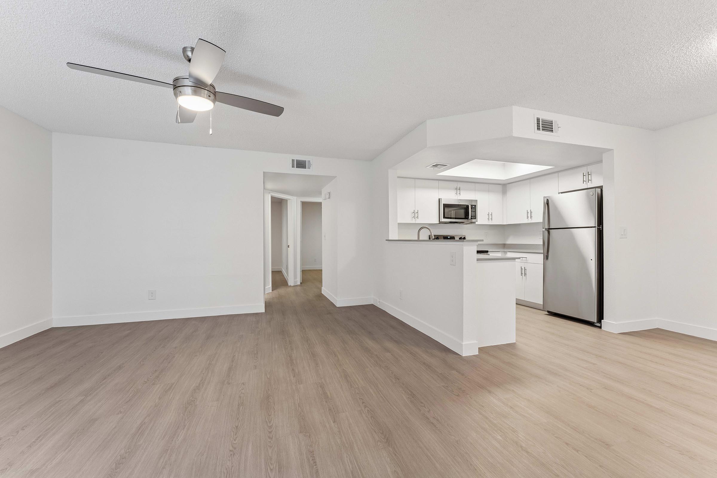 Bright and spacious living area featuring light-colored walls, laminate flooring, and a ceiling fan. An open kitchen with modern appliances, including a stainless steel refrigerator and microwave, is visible in the background. A hallway leads to additional rooms, enhancing the sense of space.