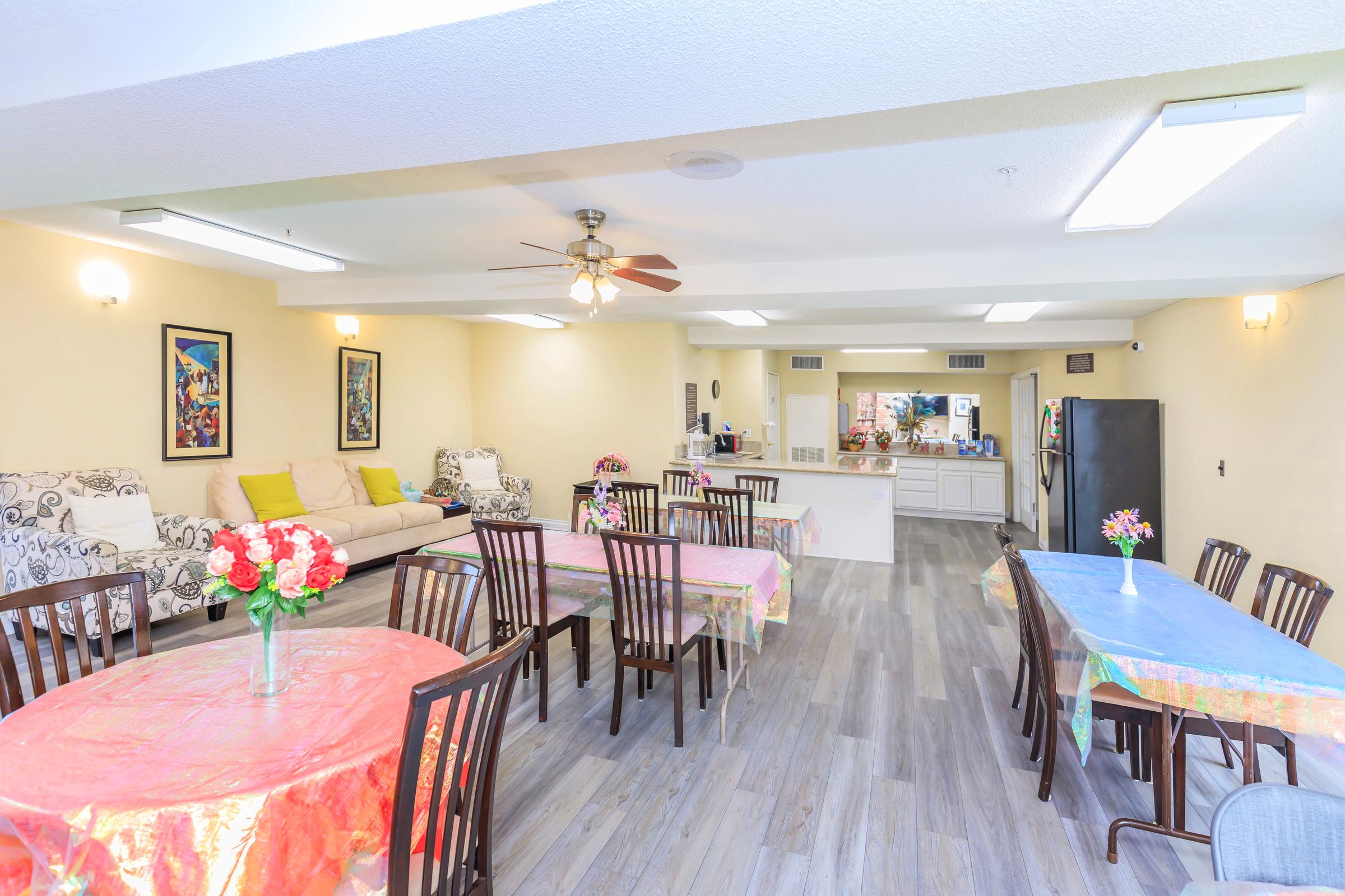 A spacious community room featuring several tables covered with colorful tablecloths, chairs, and decorative flowers. A cozy seating area with a sofa is visible, along with a kitchen area in the background. The walls are adorned with framed artwork, creating a welcoming atmosphere.