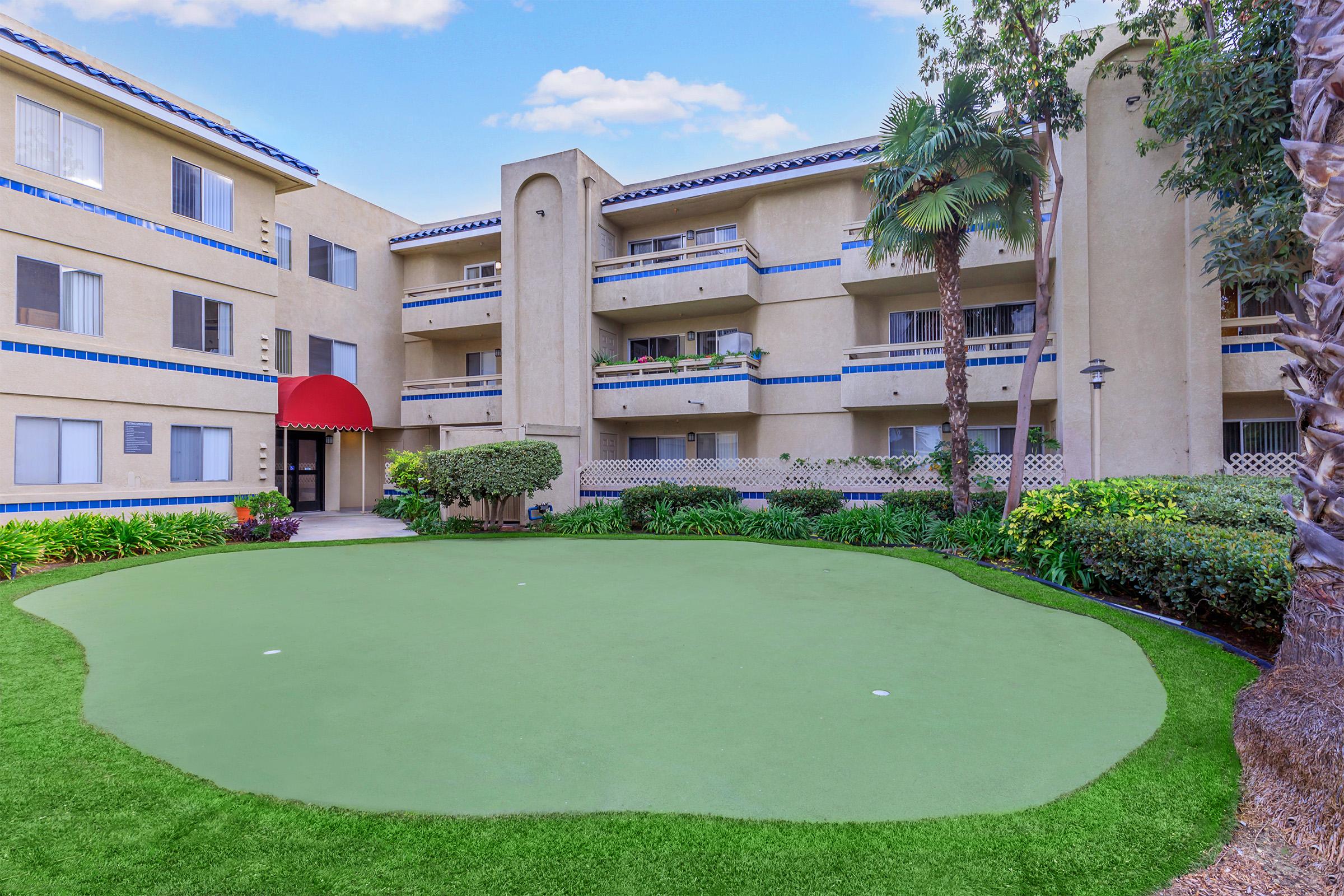 A view of a well-maintained apartment complex featuring a green lawn and a putting green. The building has multiple stories with balconies, tropical plants, and an inviting red entrance. Bright blue skies create a cheerful atmosphere in the background.