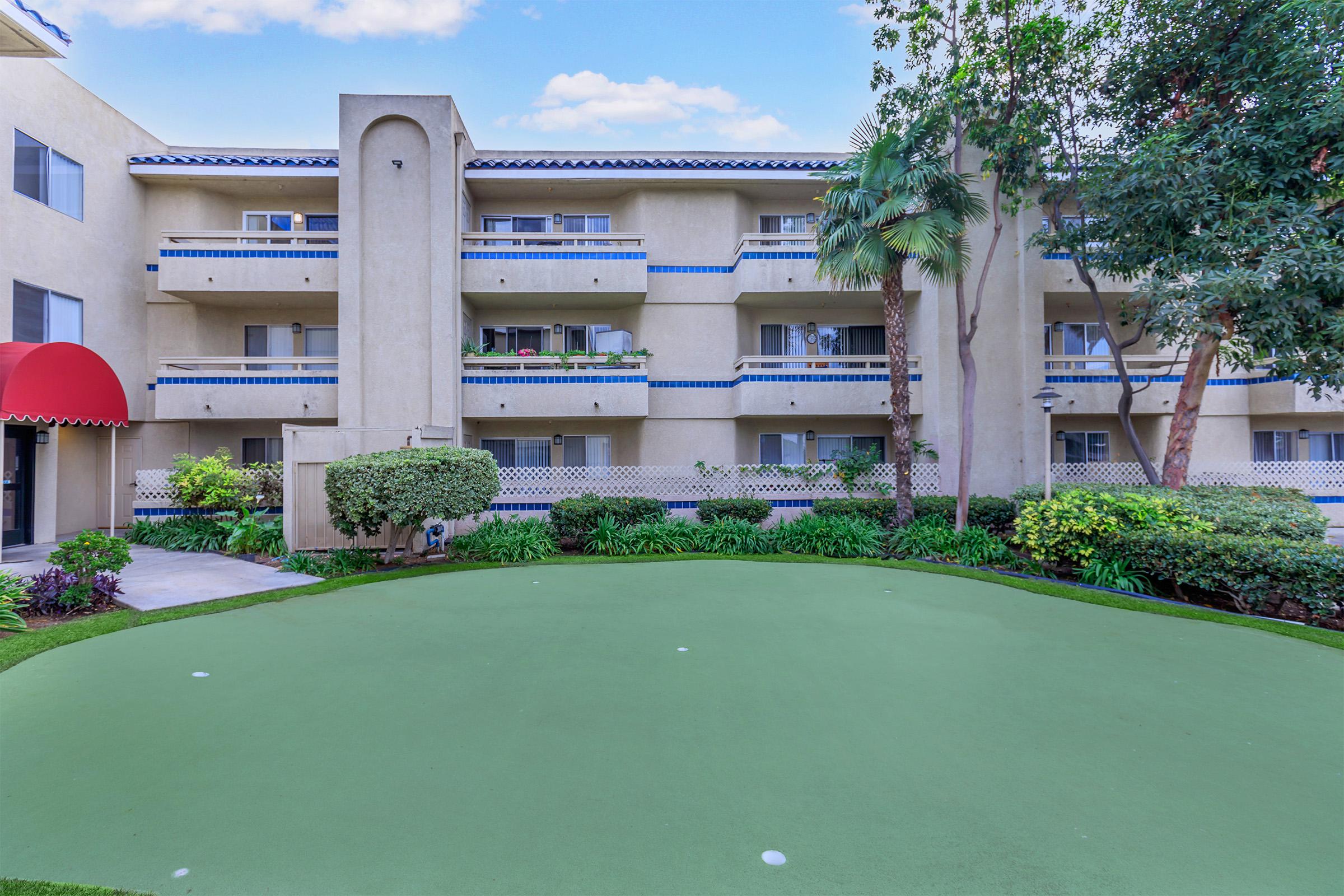 A well-maintained putting green in the foreground, surrounded by lush greenery and shrubs. In the background, a light-colored building with balconies and red awning features. The scene is bright and inviting, showcasing a tranquil outdoor space.