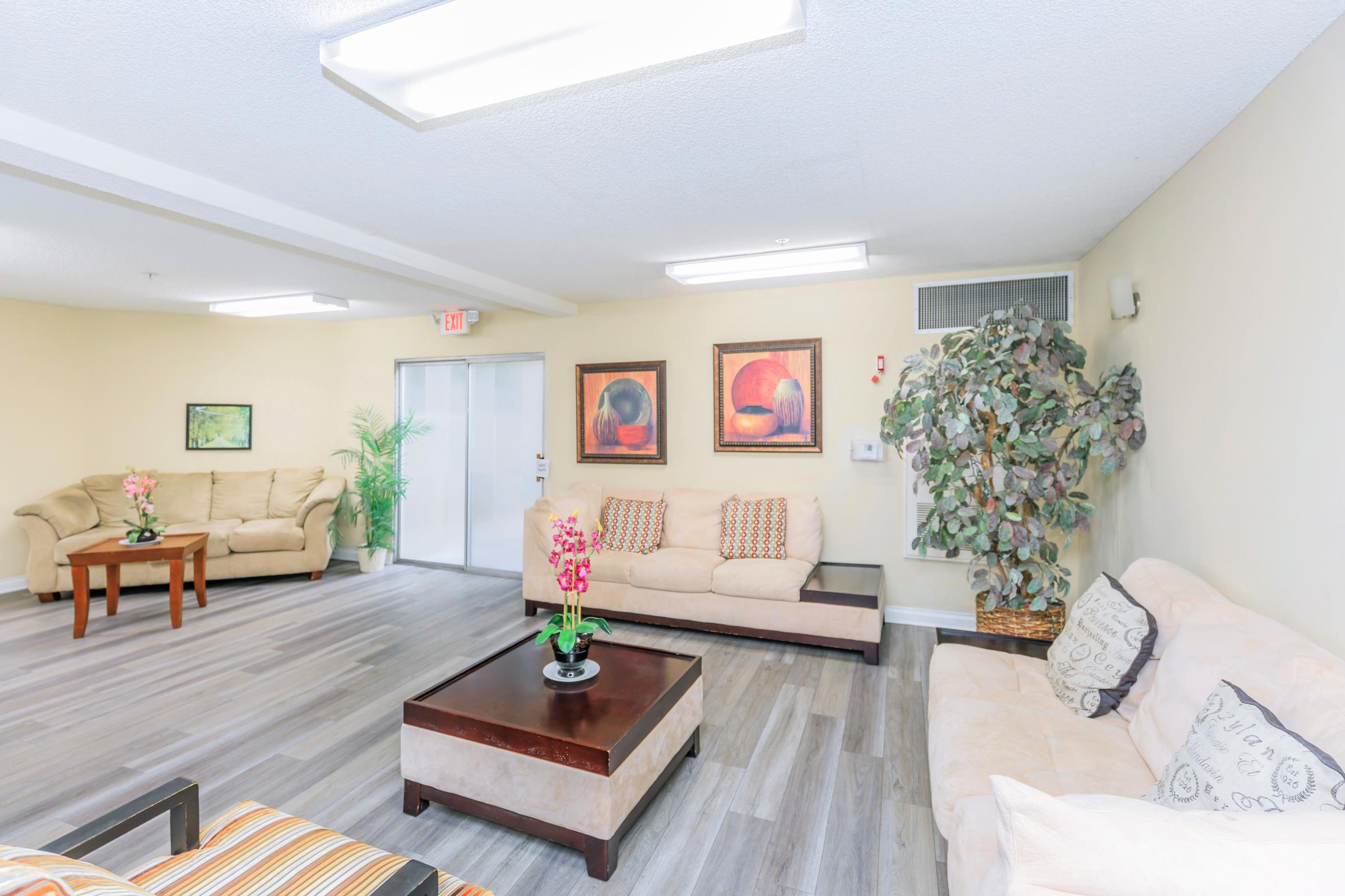 A cozy lounge area with two beige sofas and a matching loveseat, arranged around a dark wooden coffee table. Decorative plants and two framed paintings adorn the walls, while a glass door adds a touch of openness. Natural light from ceiling fixtures enhances the inviting atmosphere.