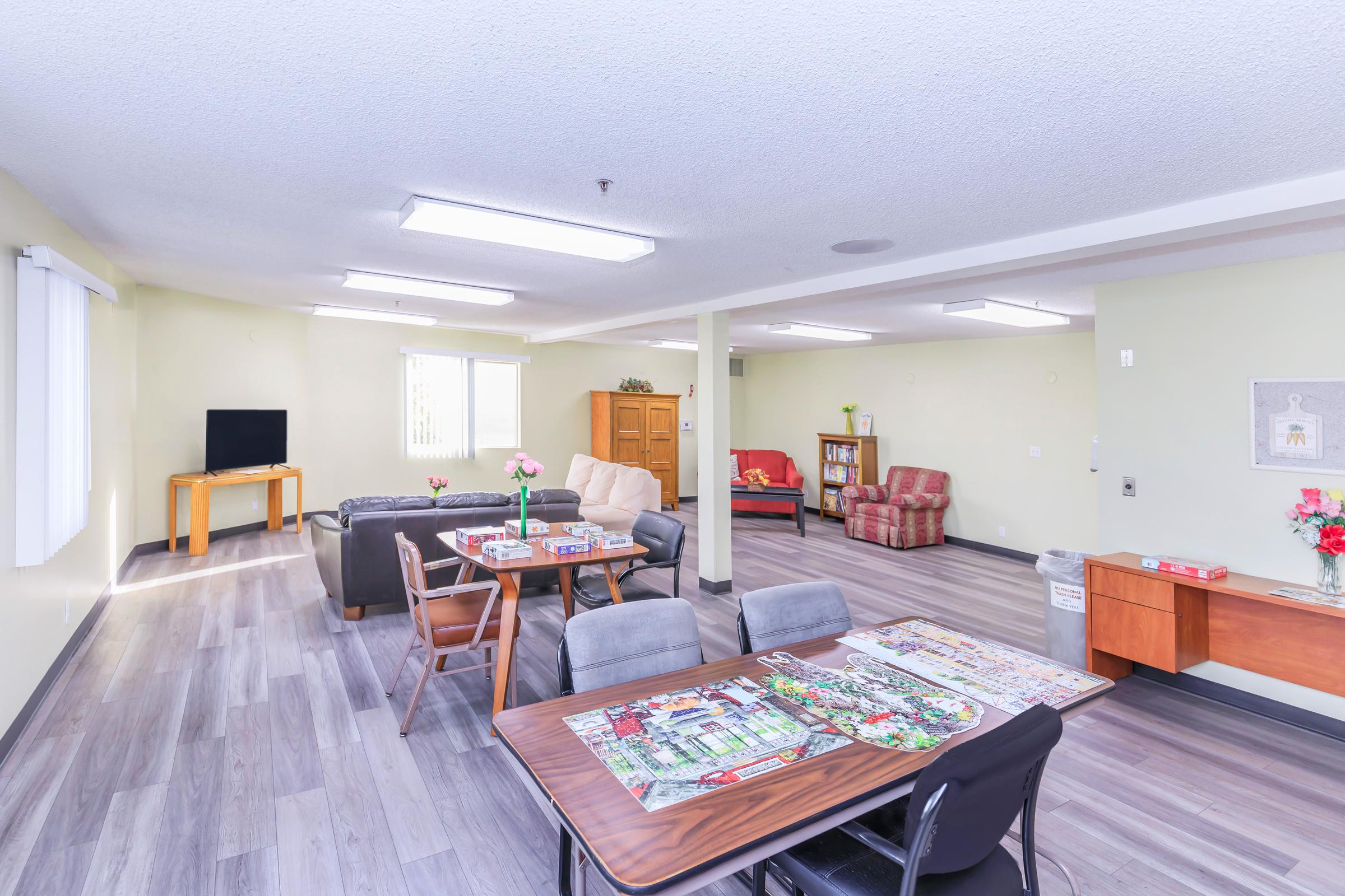 A spacious common area featuring a seating arrangement with couches, armchairs, and a television. There are tables set up for activities, decorated with puzzles and flowers. The walls are light-colored, and there are windows allowing natural light. A cabinet and a trash bin are visible, contributing to a welcoming environment.