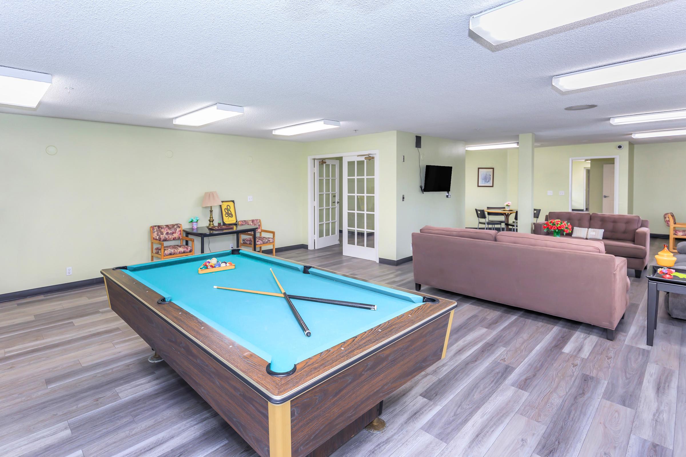 A well-lit game room featuring a billiards table with cues and balls, plush seating areas including two chairs and a sofa, a small table with a lamp, and a door leading to another room. The walls are painted a light green, and the flooring is a light wood laminate.